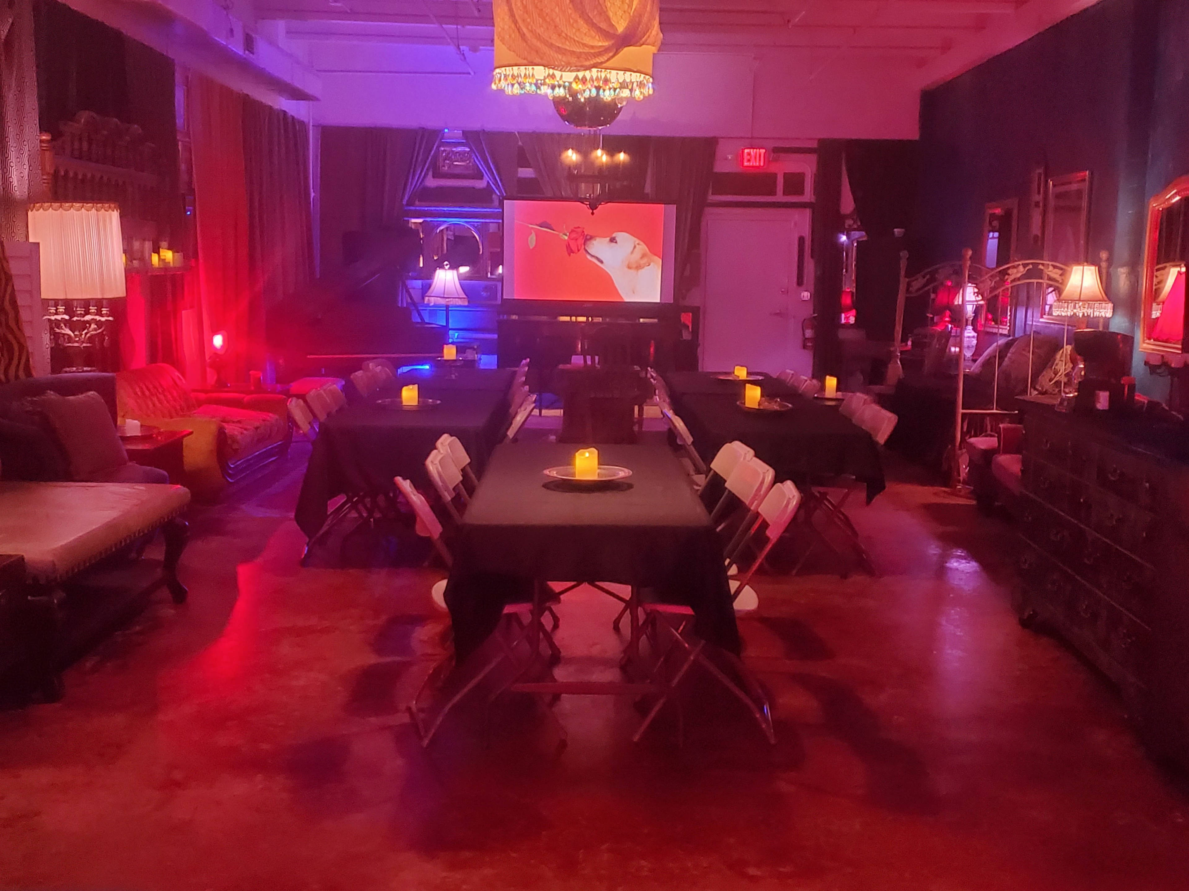 A dimly lit room with multiple tables covered in black cloth, surrounded by chairs, and illuminated by soft colored lights and candles, with a projector screen displaying an image at the far end.