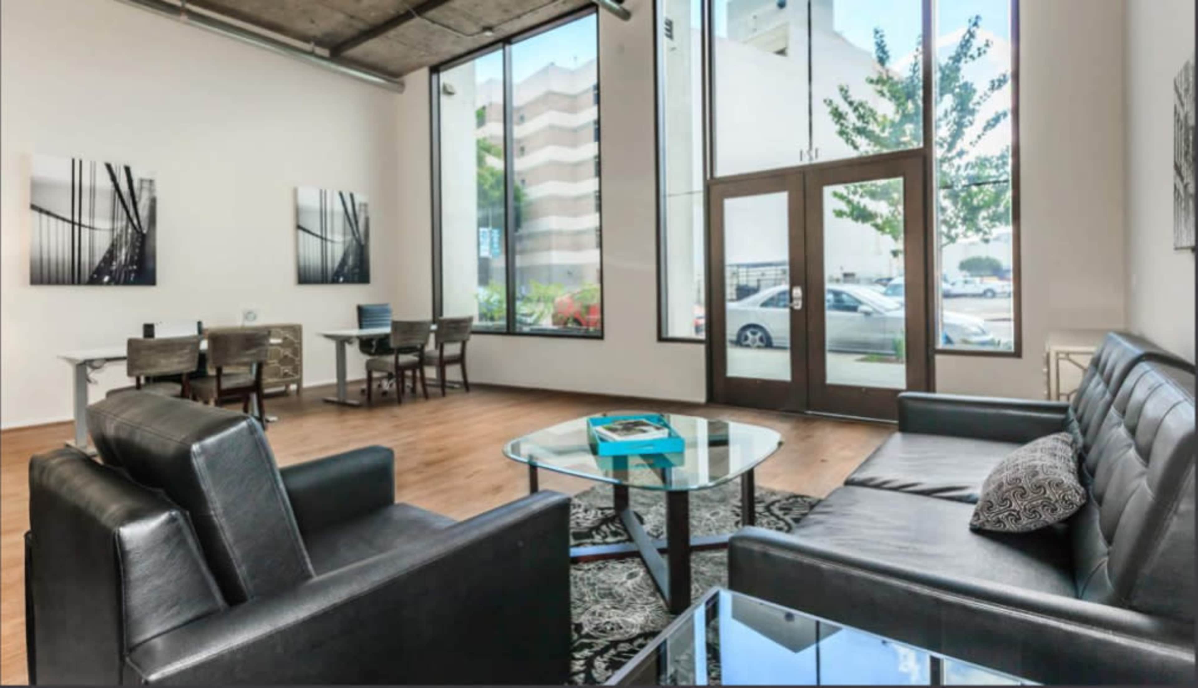 The image shows a modern interior space with large windows, a seating area featuring black leather sofas, a glass coffee table, and a dining area with a table and chairs.