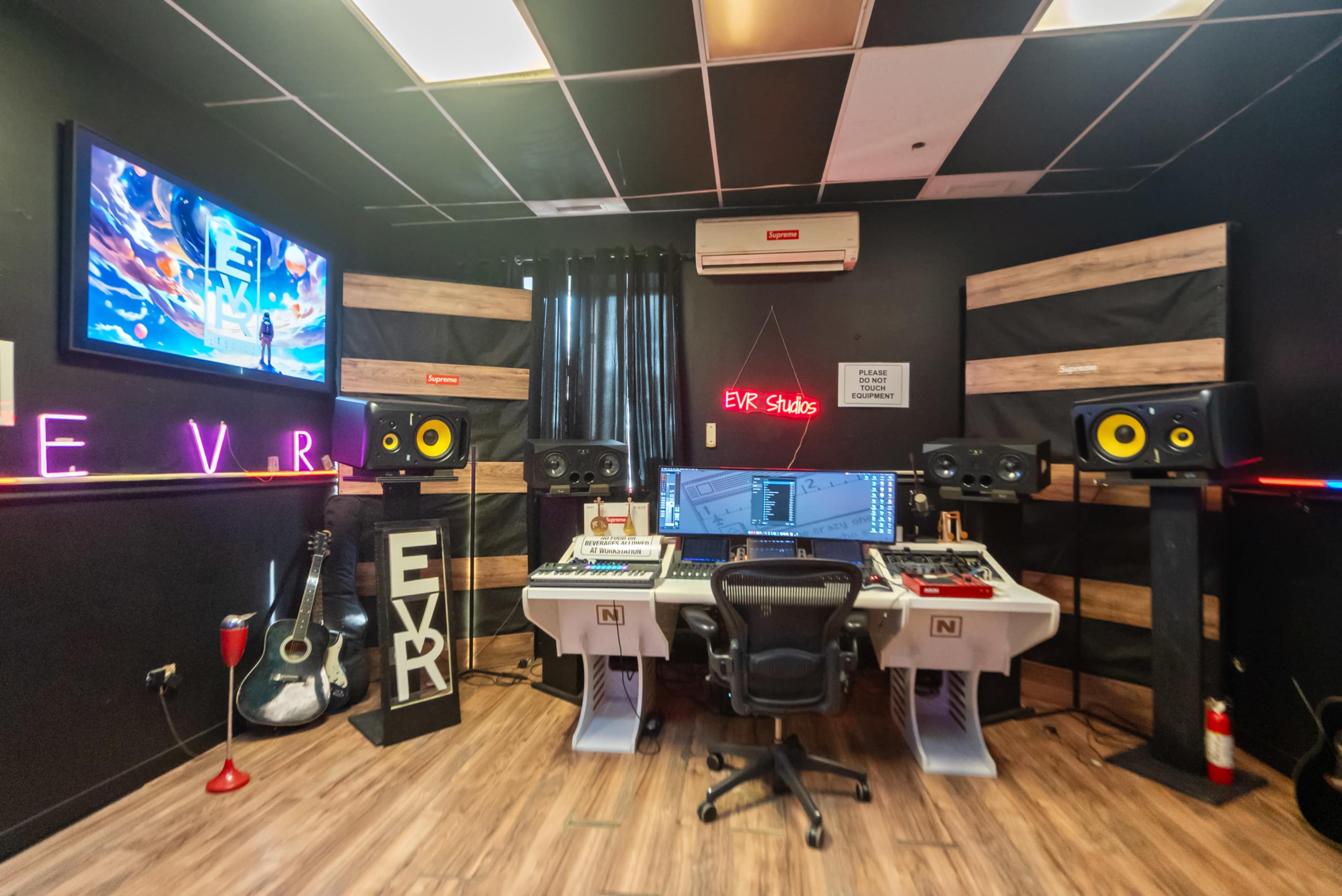 The image shows a music studio with a mixing desk, multiple speakers, a computer setup, and various musical instruments, all set against dark walls and wooden accents.