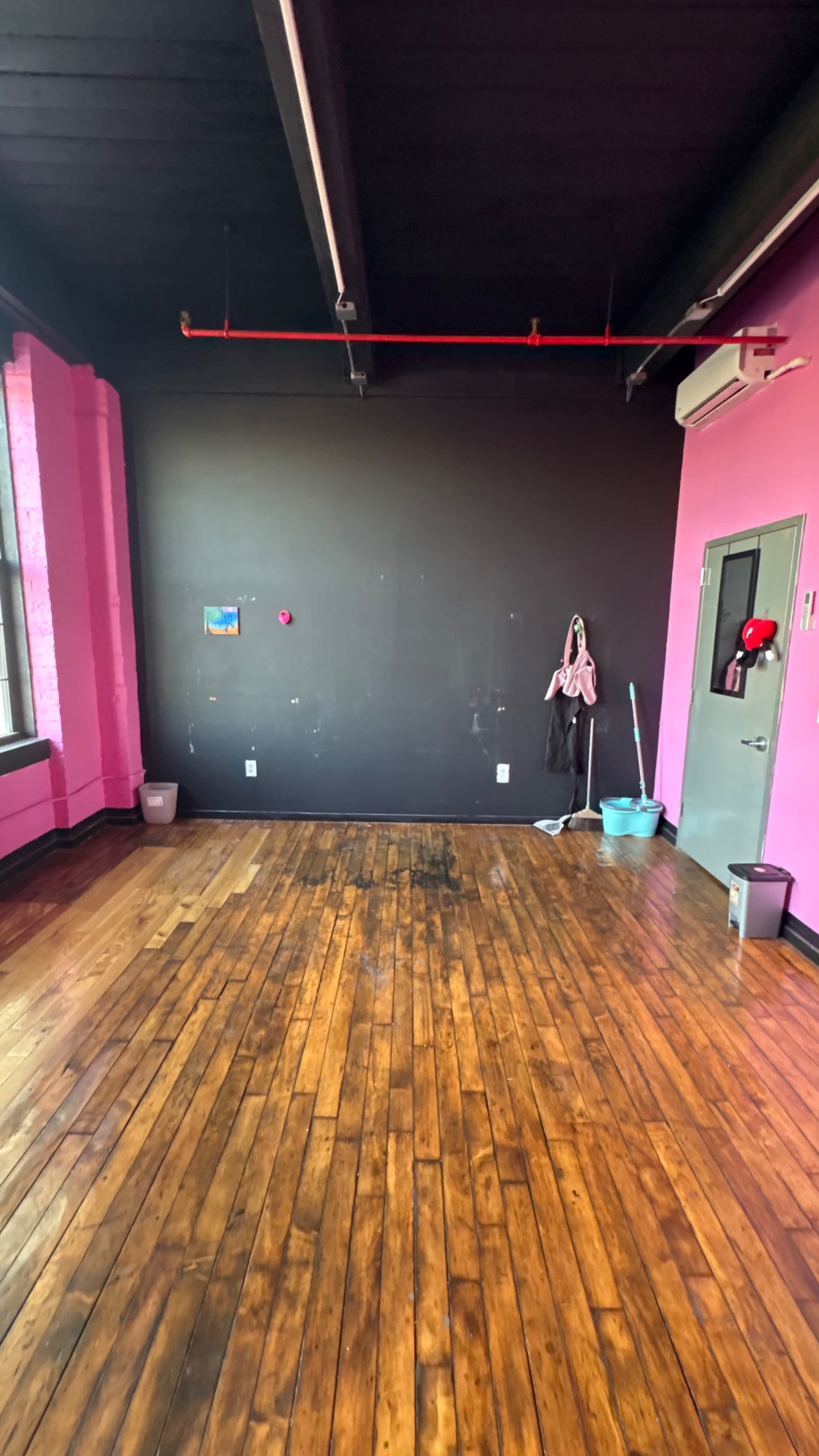 The image shows a spacious room with wooden flooring, black and pink walls, and cleaning supplies in the corners.