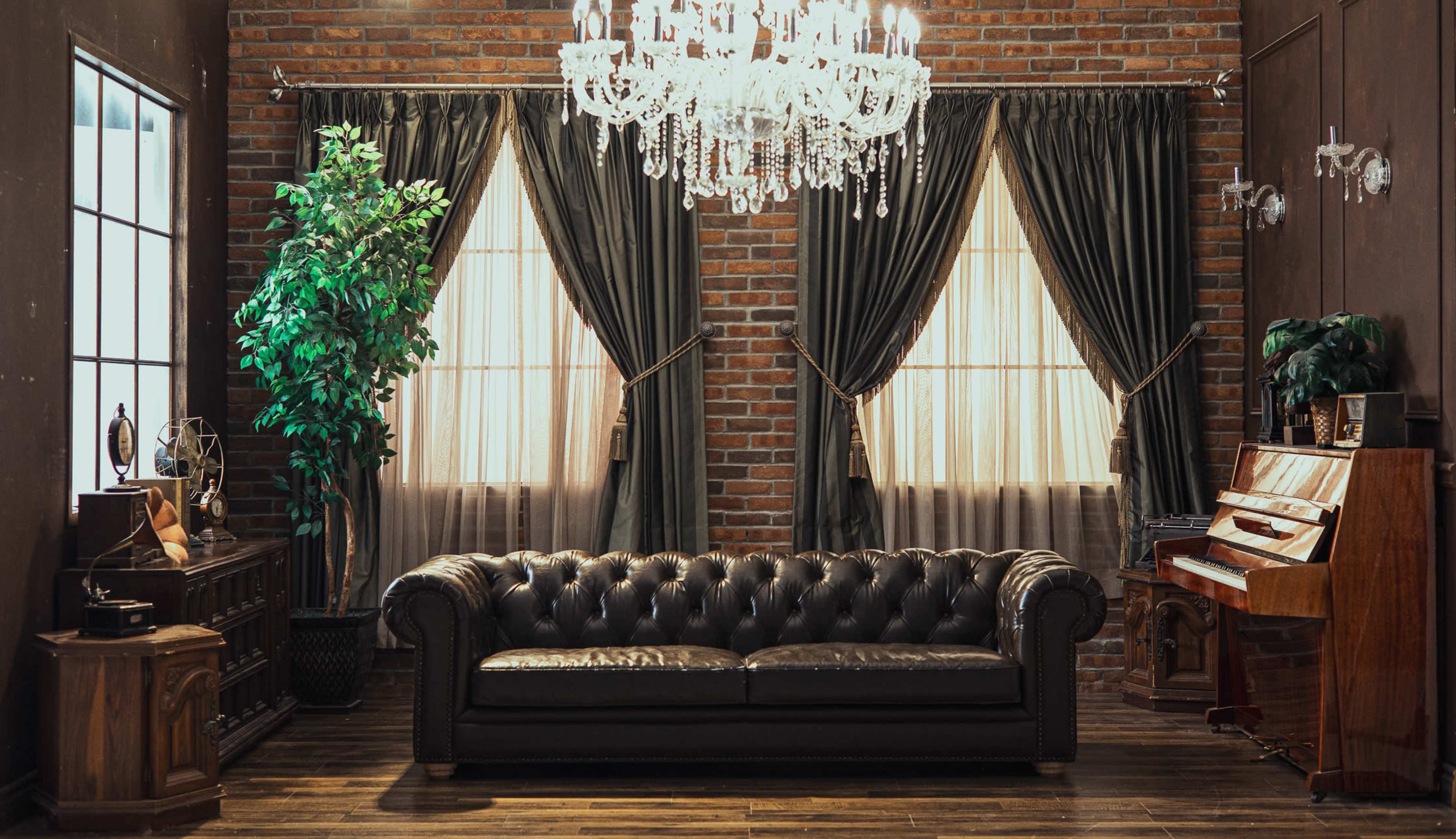 The image features an ornate room with a black leather tufted sofa, a crystal chandelier, and large windows draped with dark curtains, bordered by brick walls.