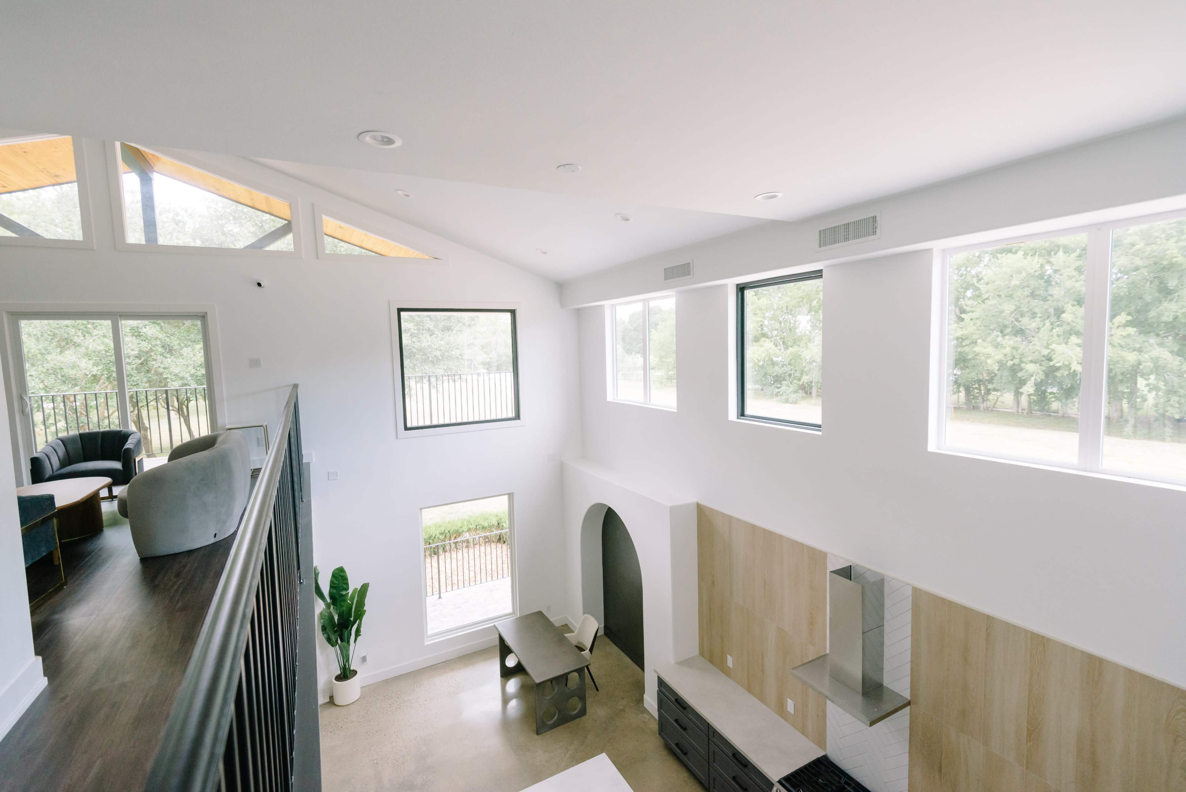 The image shows a modern living space with large windows, a high ceiling, and a view of greenery outside.