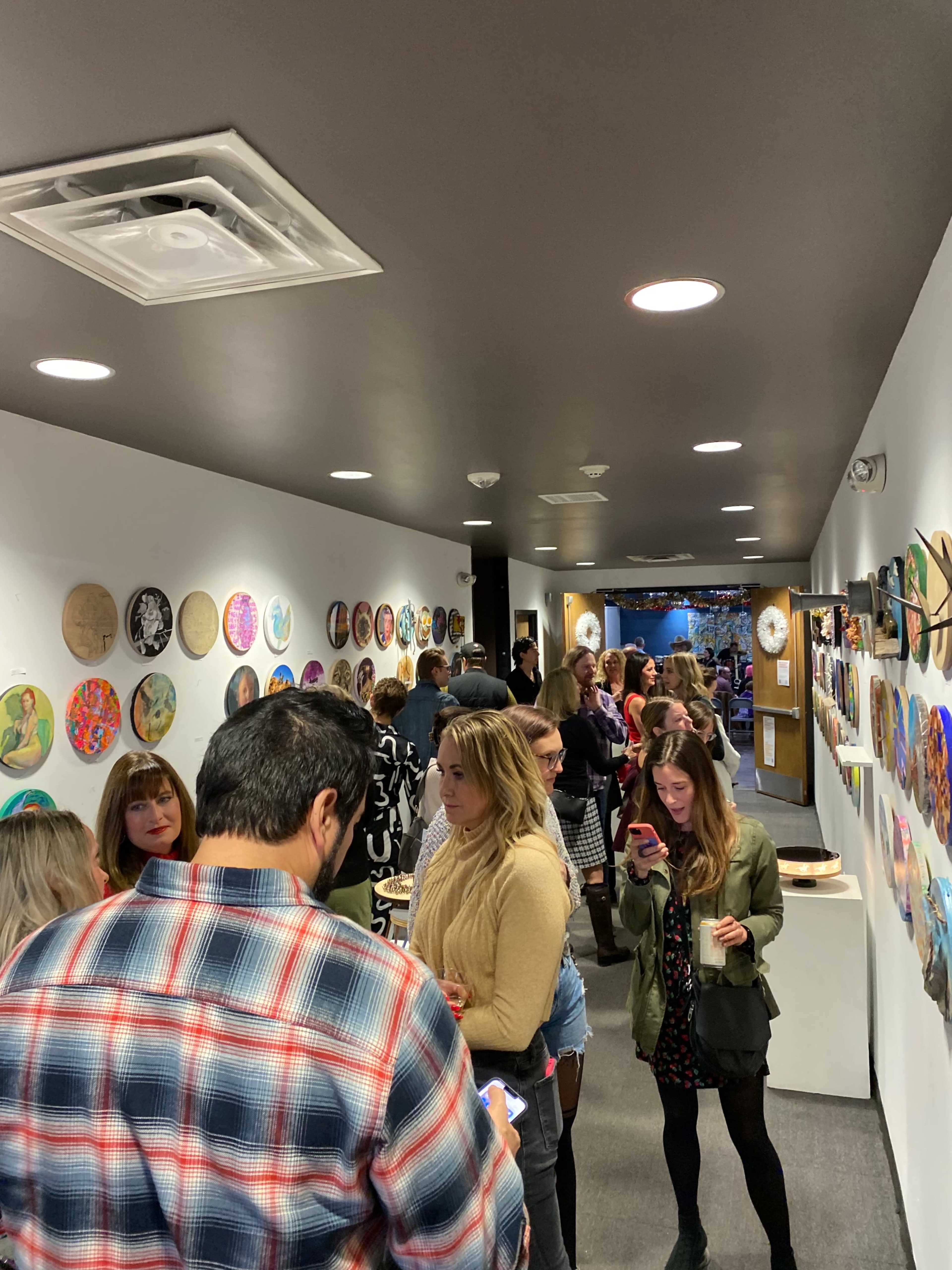 A crowded hallway features people engaging with artwork displayed on the walls.