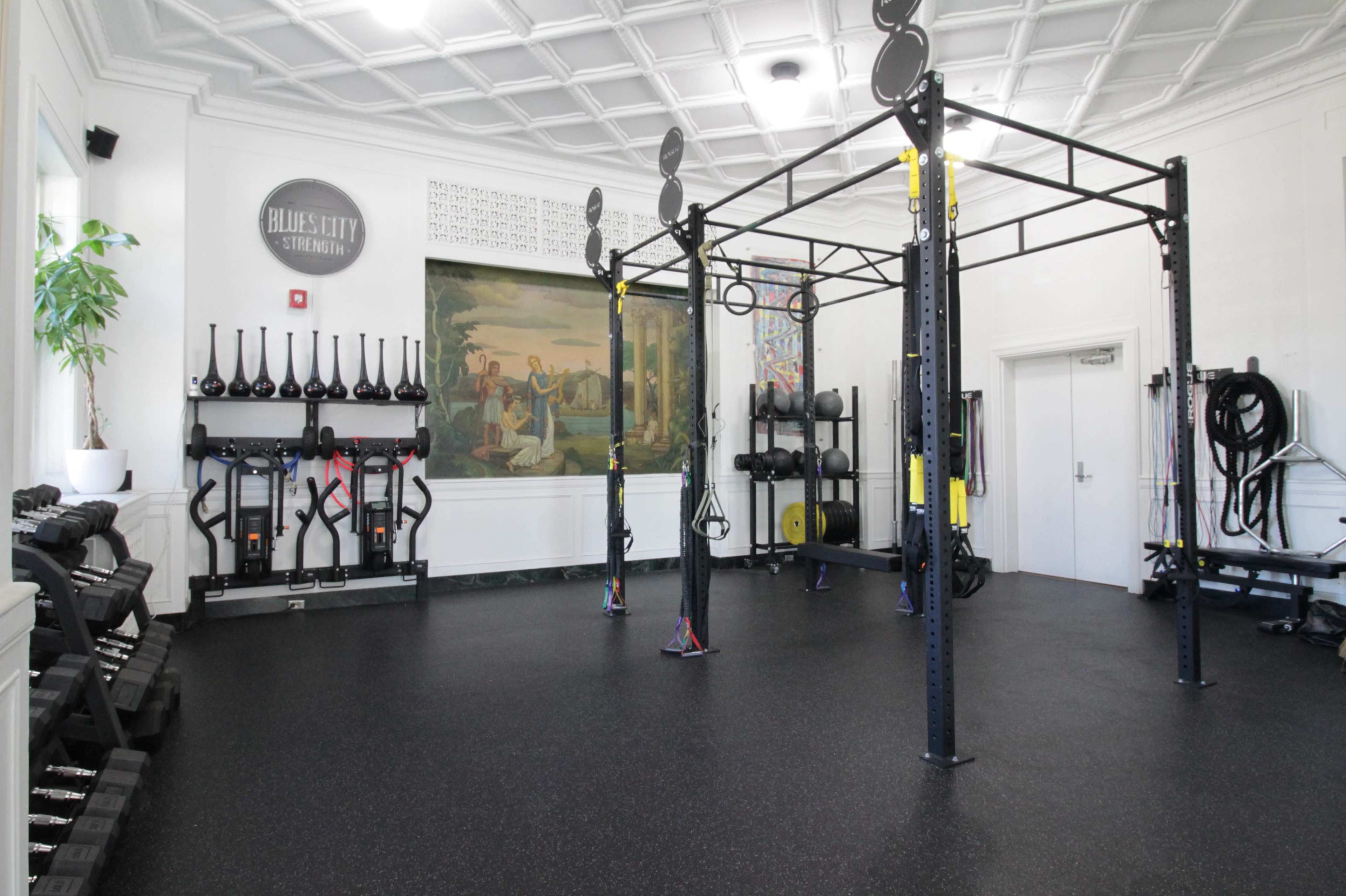 The image shows a modern fitness area equipped with various exercise equipment, including dumbbells, resistance bands, and a multi-use training rack against a backdrop of decorative murals.