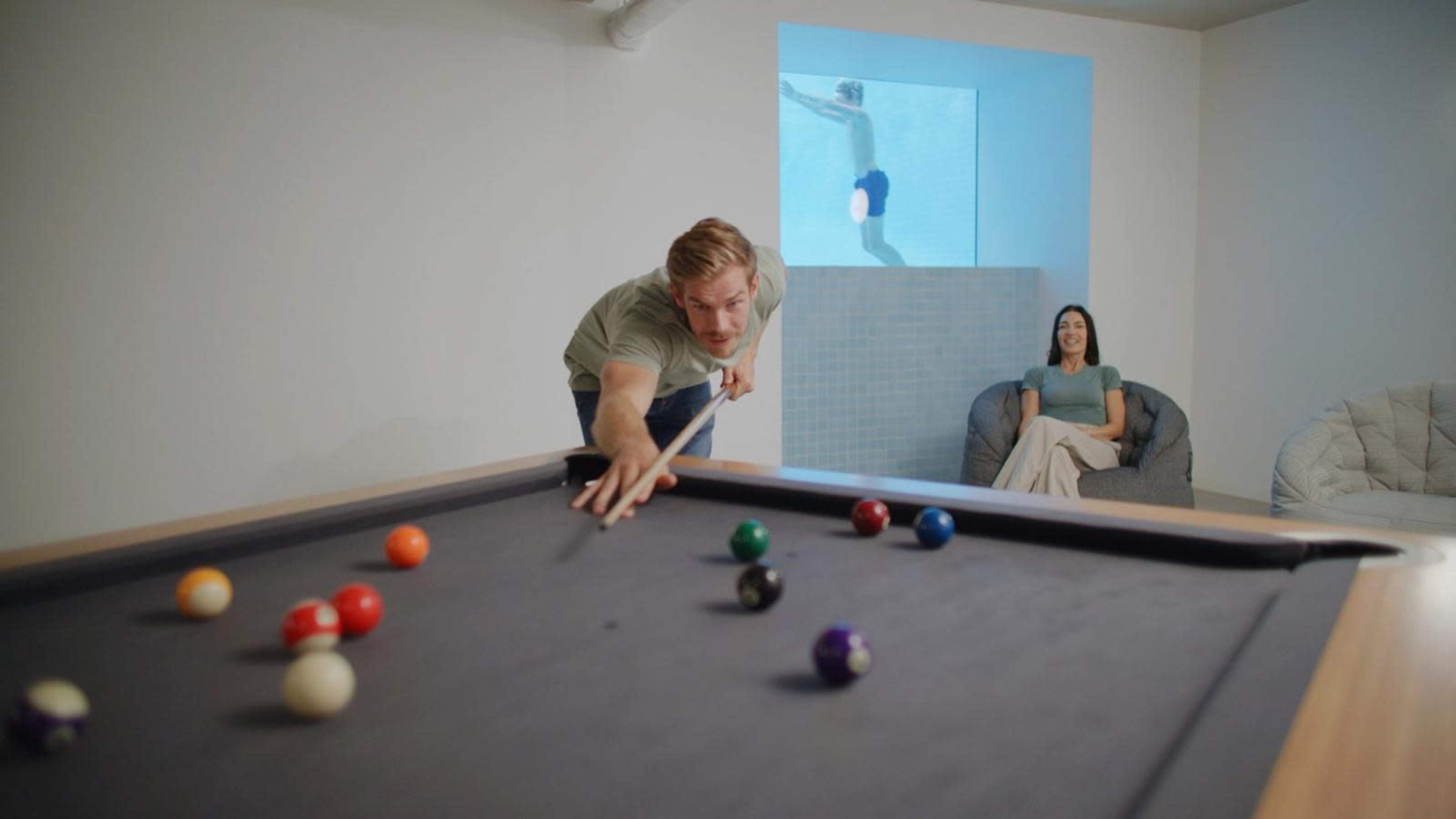 A man leans over a pool table while a woman sits on a couch, and a person swims in a pool visible through a window.