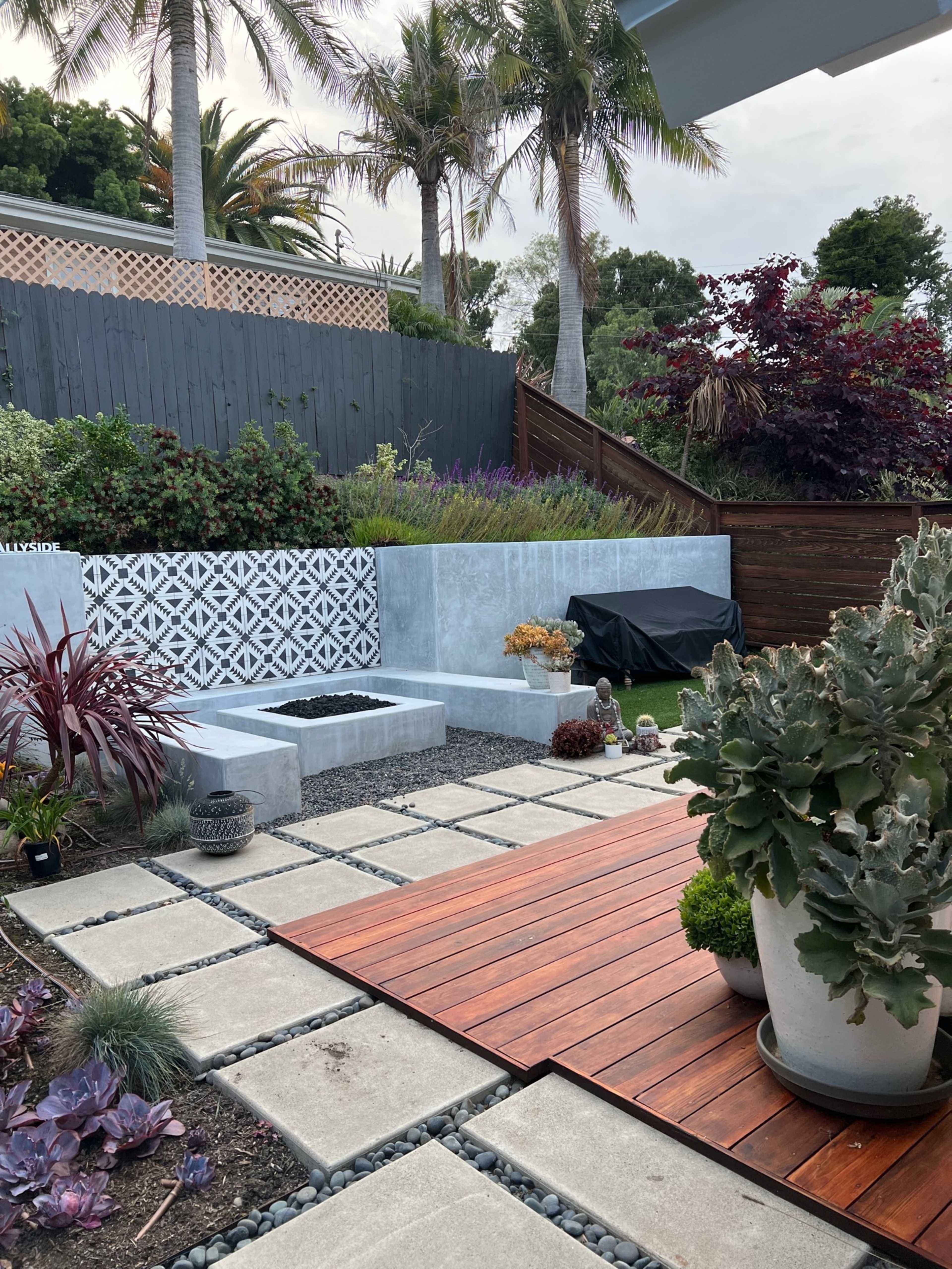 A landscaped outdoor space featuring stone seating, wooden decking, various plants, and a tiled wall, surrounded by greenery and palm trees.