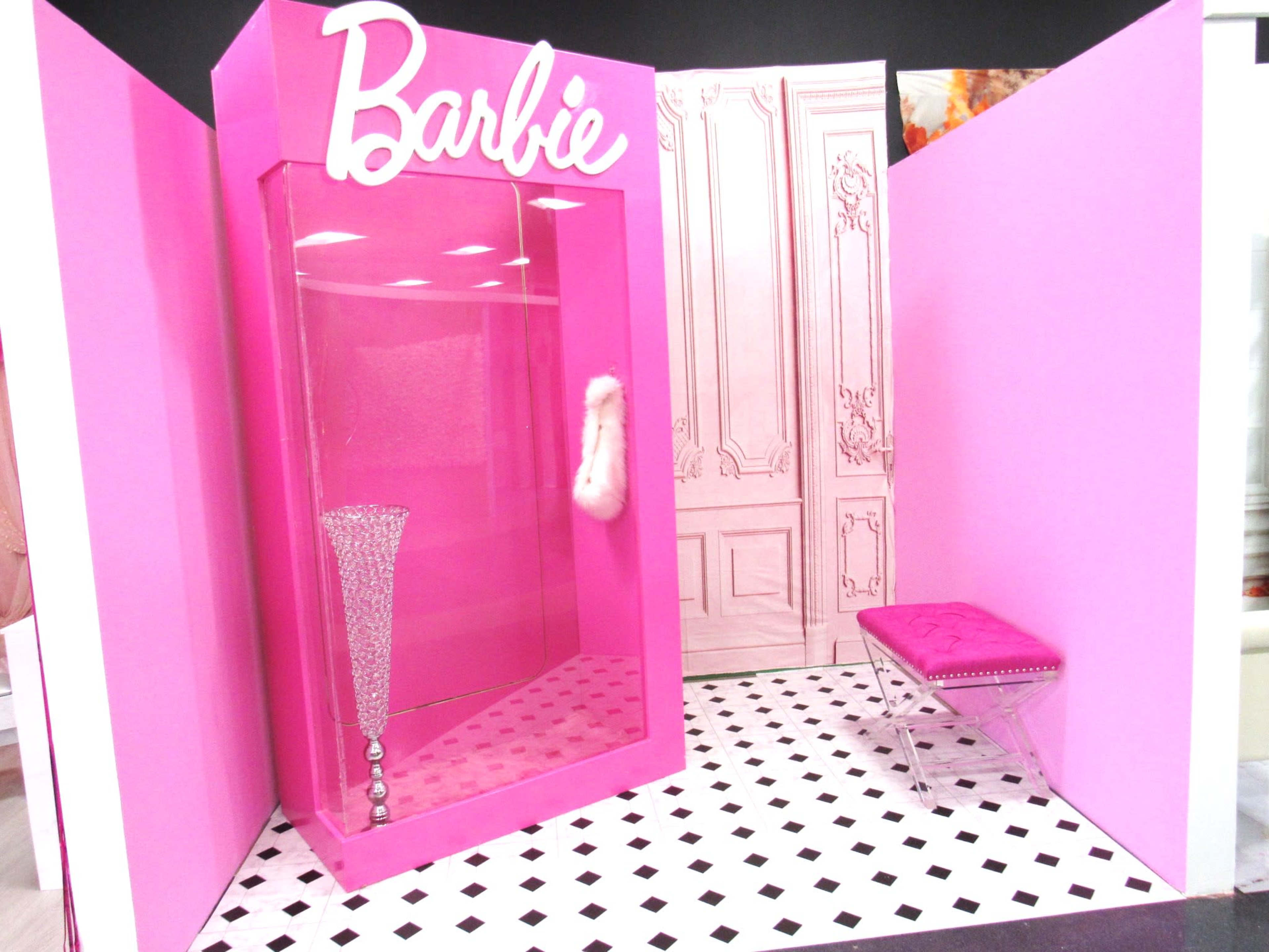 A pink display featuring a glass case labeled "Barbie," with a crystal vase, a pink fur item, and a clear acrylic bench in a room with a patterned floor.