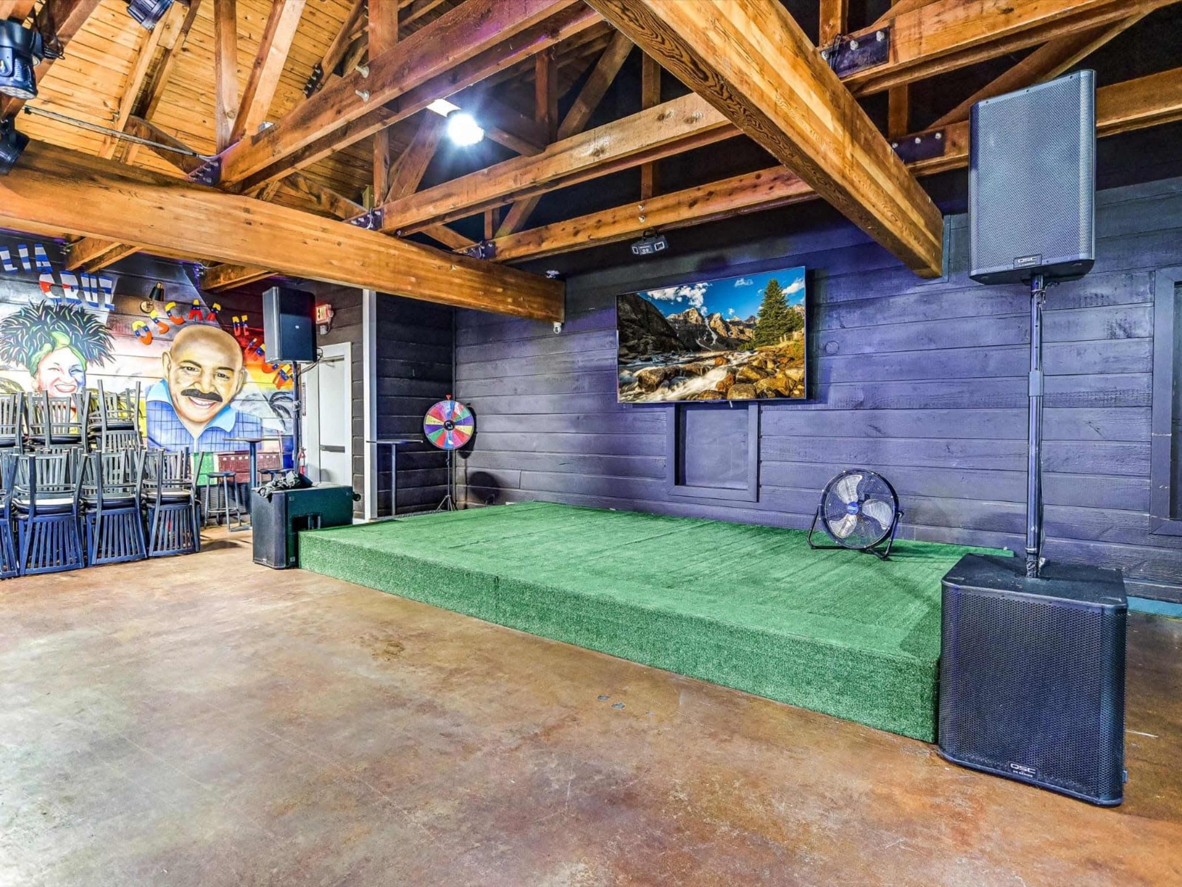 The image shows a performance stage with green carpeting, flanked by large speakers, in a room featuring wooden beams and a mural on one wall.