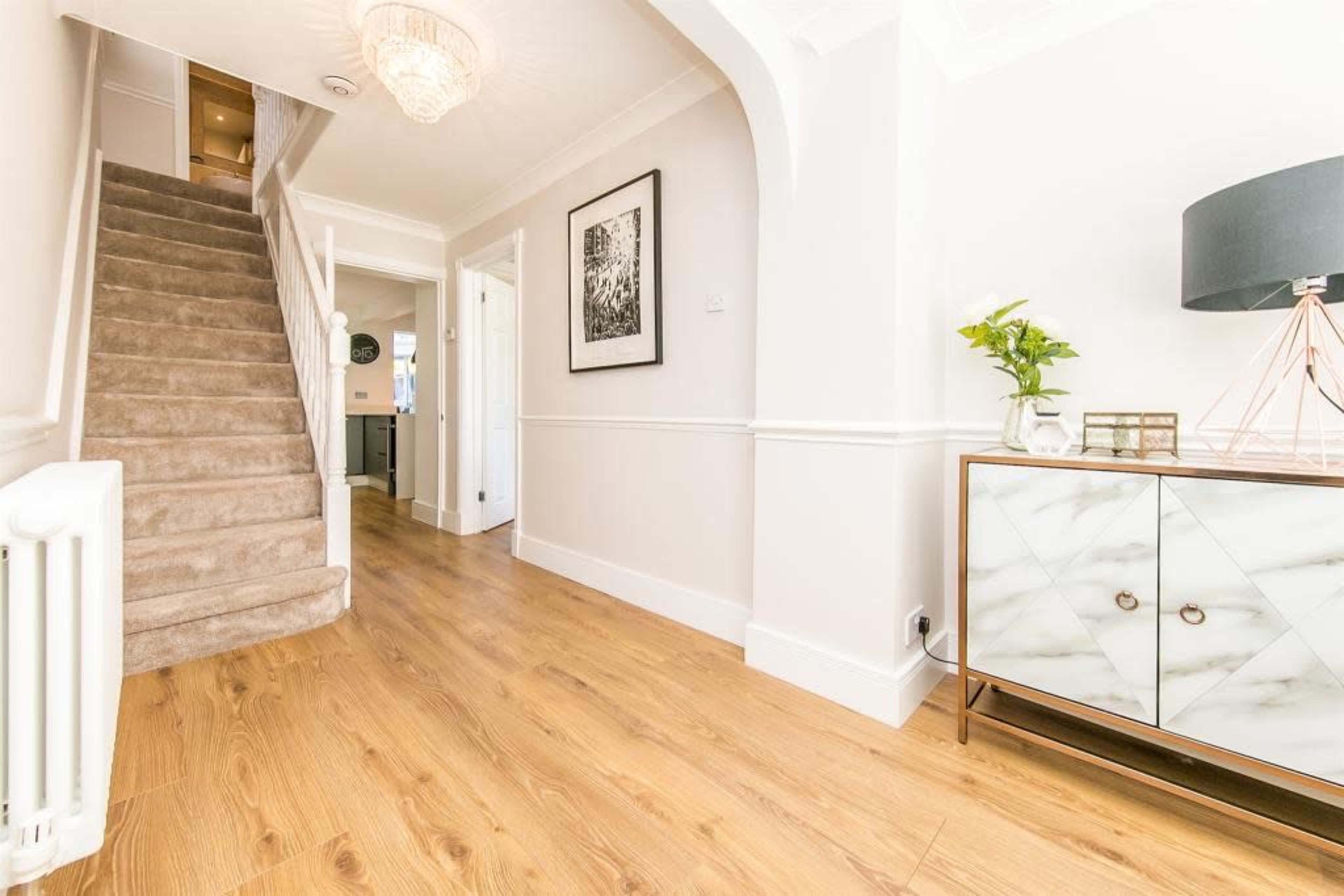 The image shows a bright hallway featuring a staircase on the left, a decorative side table on the right, and light-colored walls.