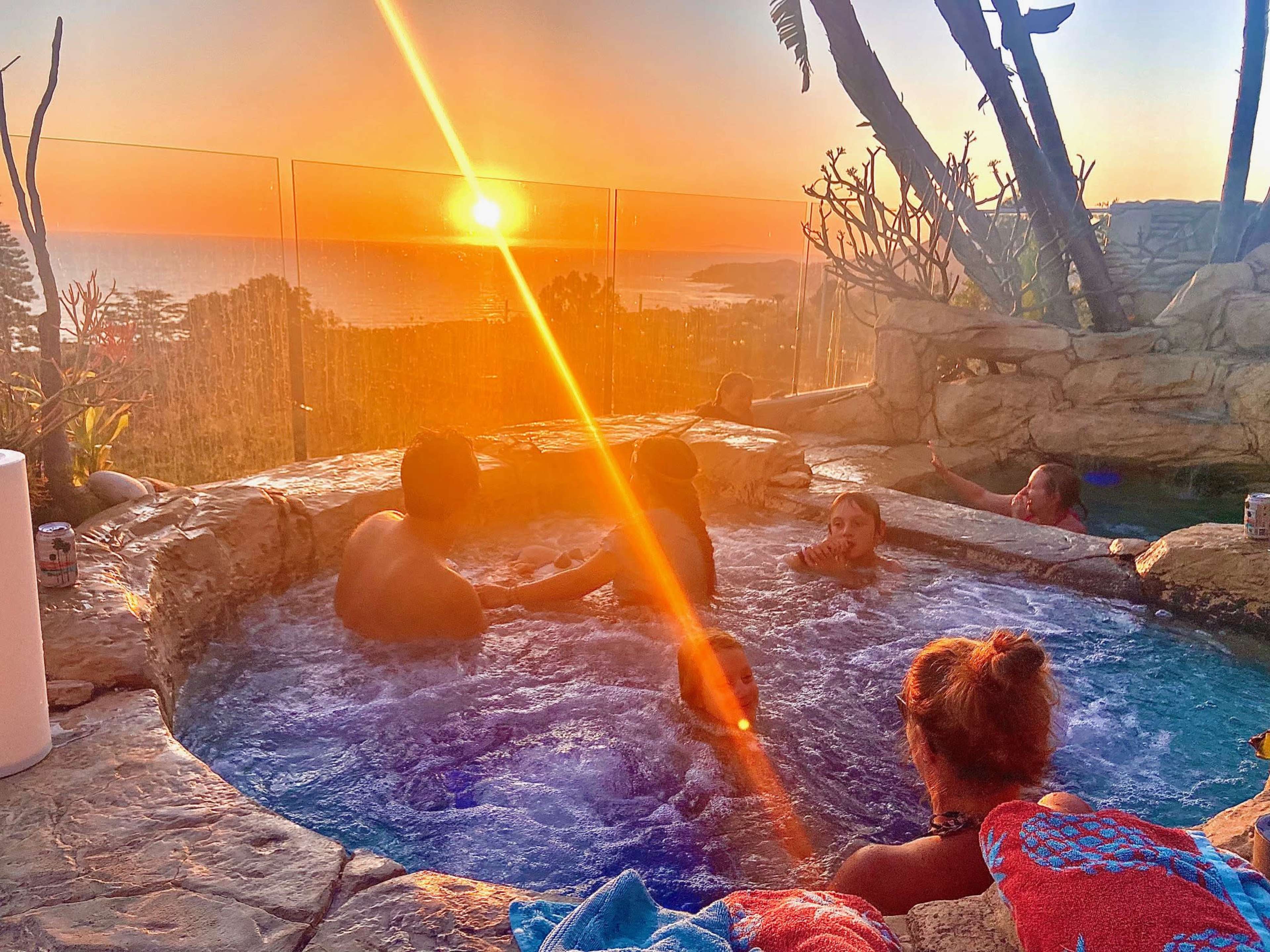 A group of people relaxes in a hot tub while watching the sun set over the ocean.