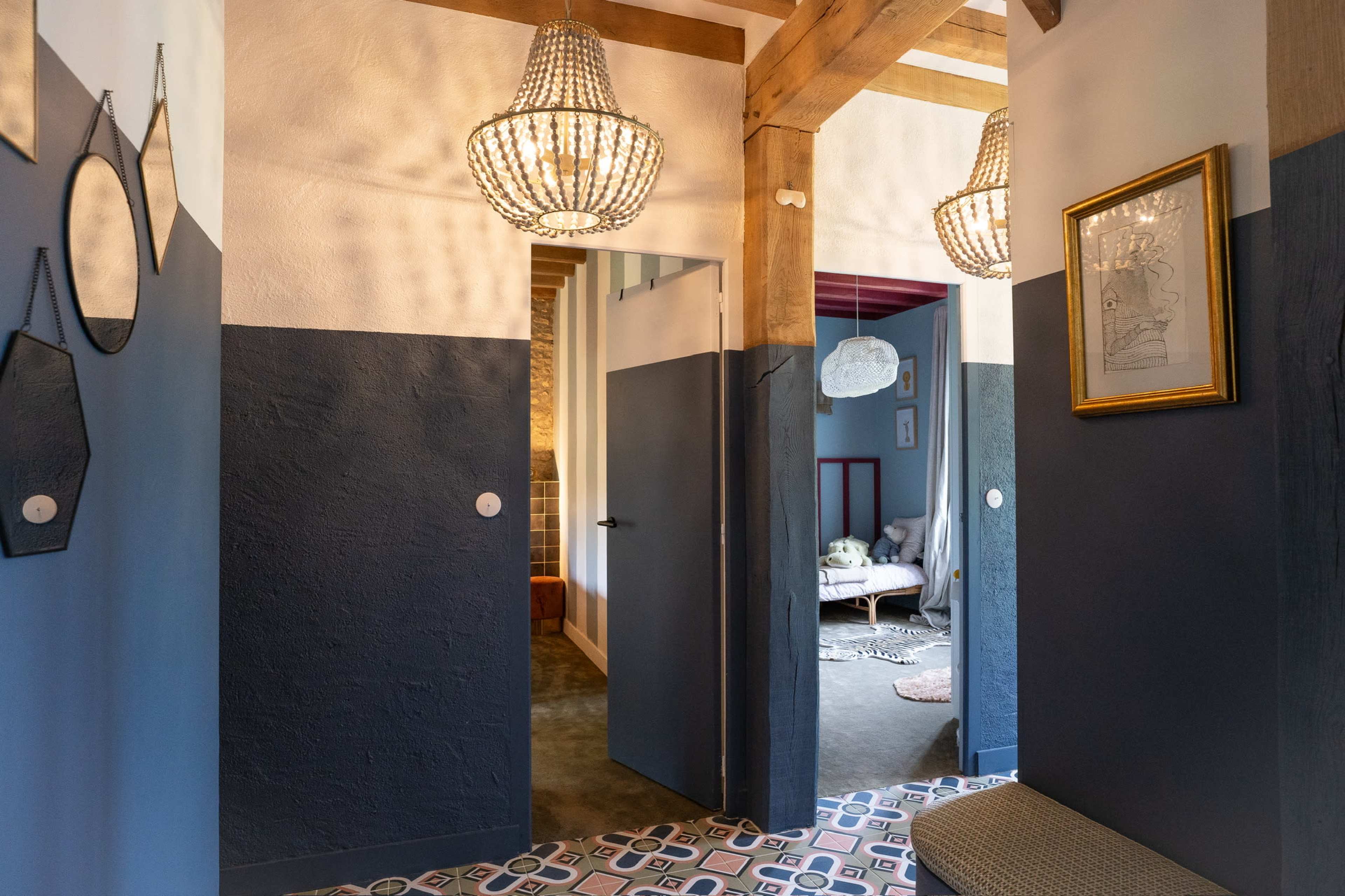 The image shows a hallway with wooden beams, two hanging chandeliers, and a patterned floor, leading to a room beyond.
