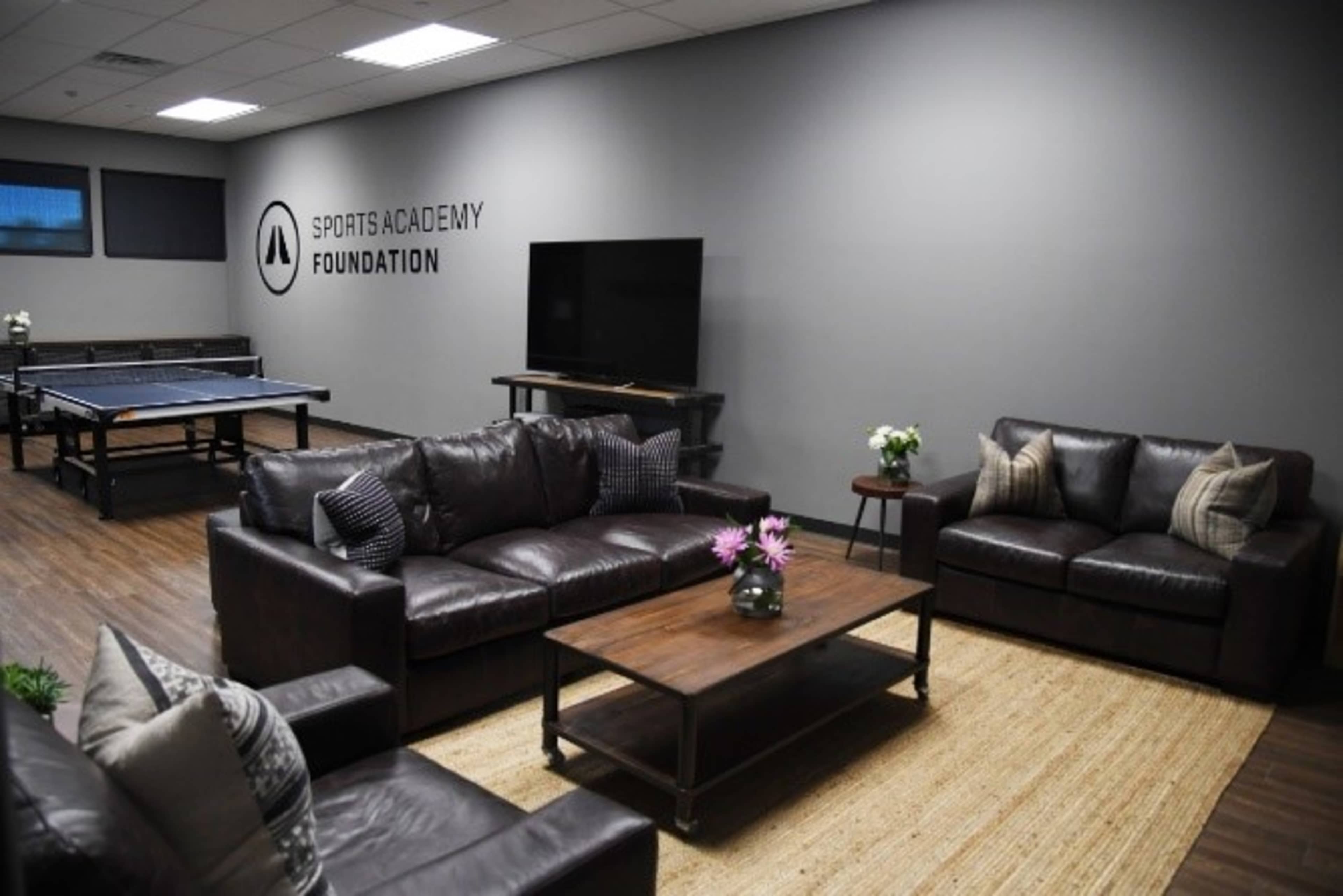 A lounge area with two leather couches, a coffee table, and a ping pong table in the background, under the logo of the Sports Academy Foundation.