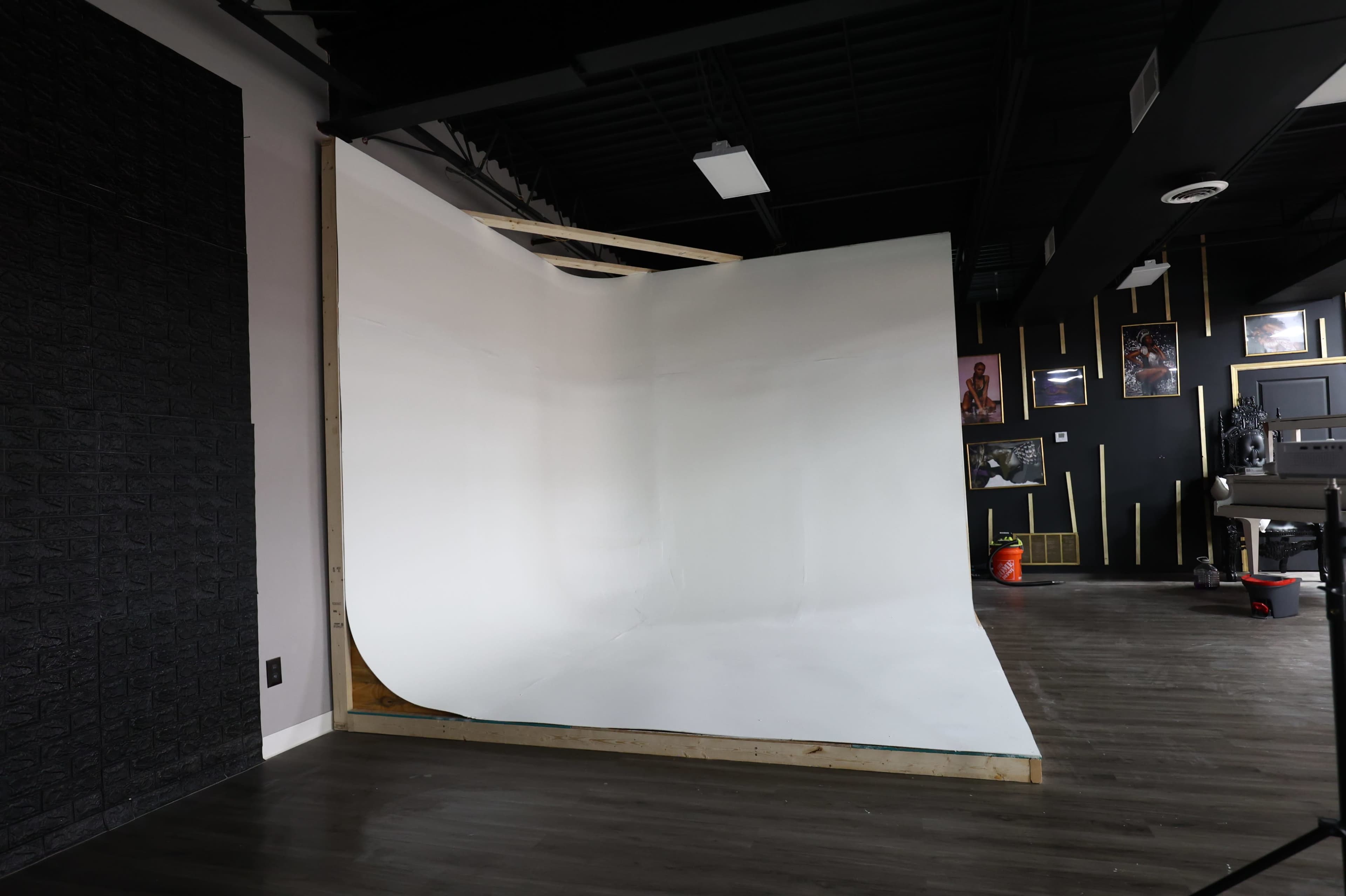 The image shows a spacious studio with a white seamless backdrop and dark wood flooring, featuring black walls with framed artwork.