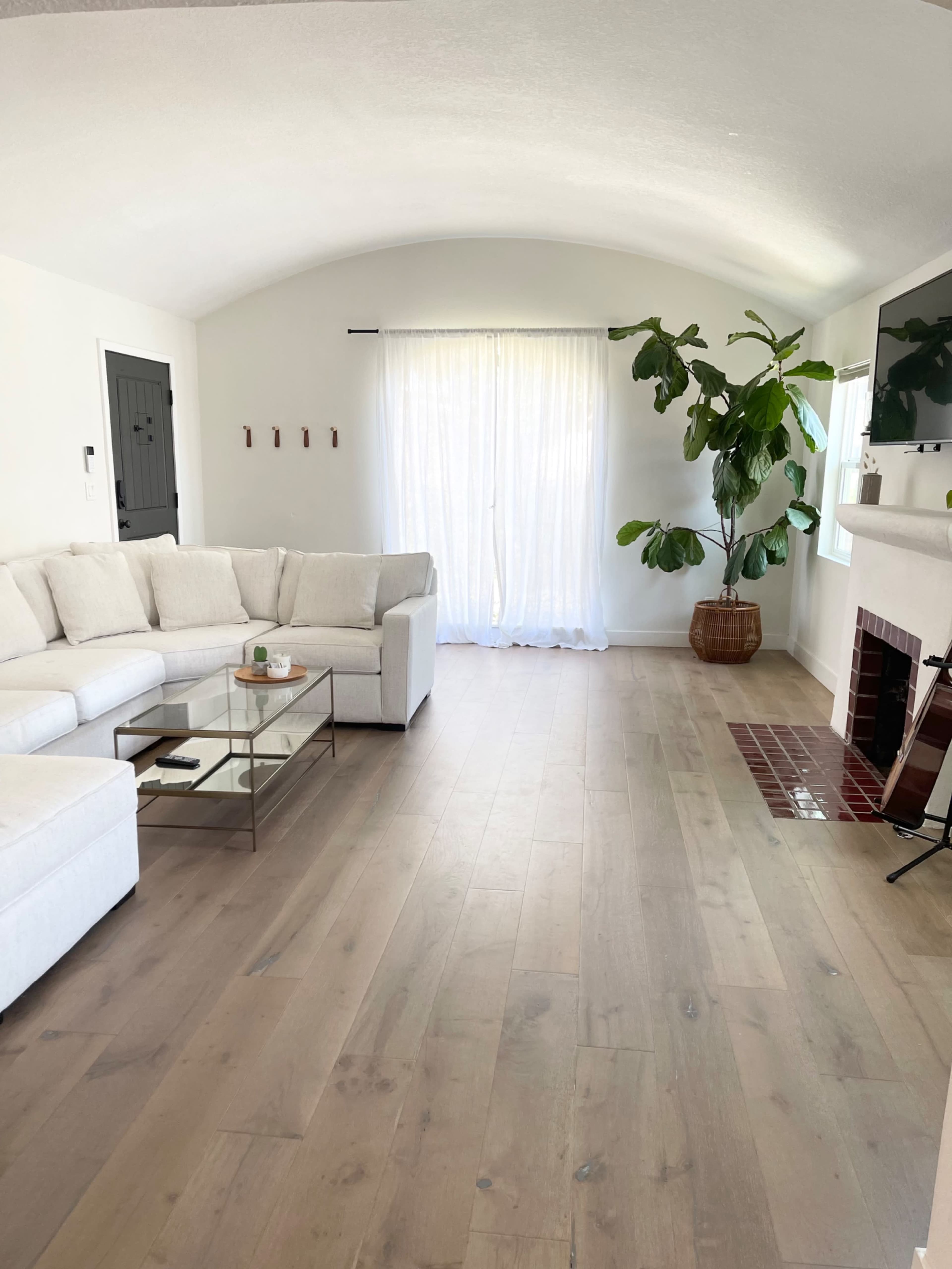 A bright living room with a white sectional sofa, a glass coffee table, large windows with sheer curtains, and a potted plant near a white fireplace.