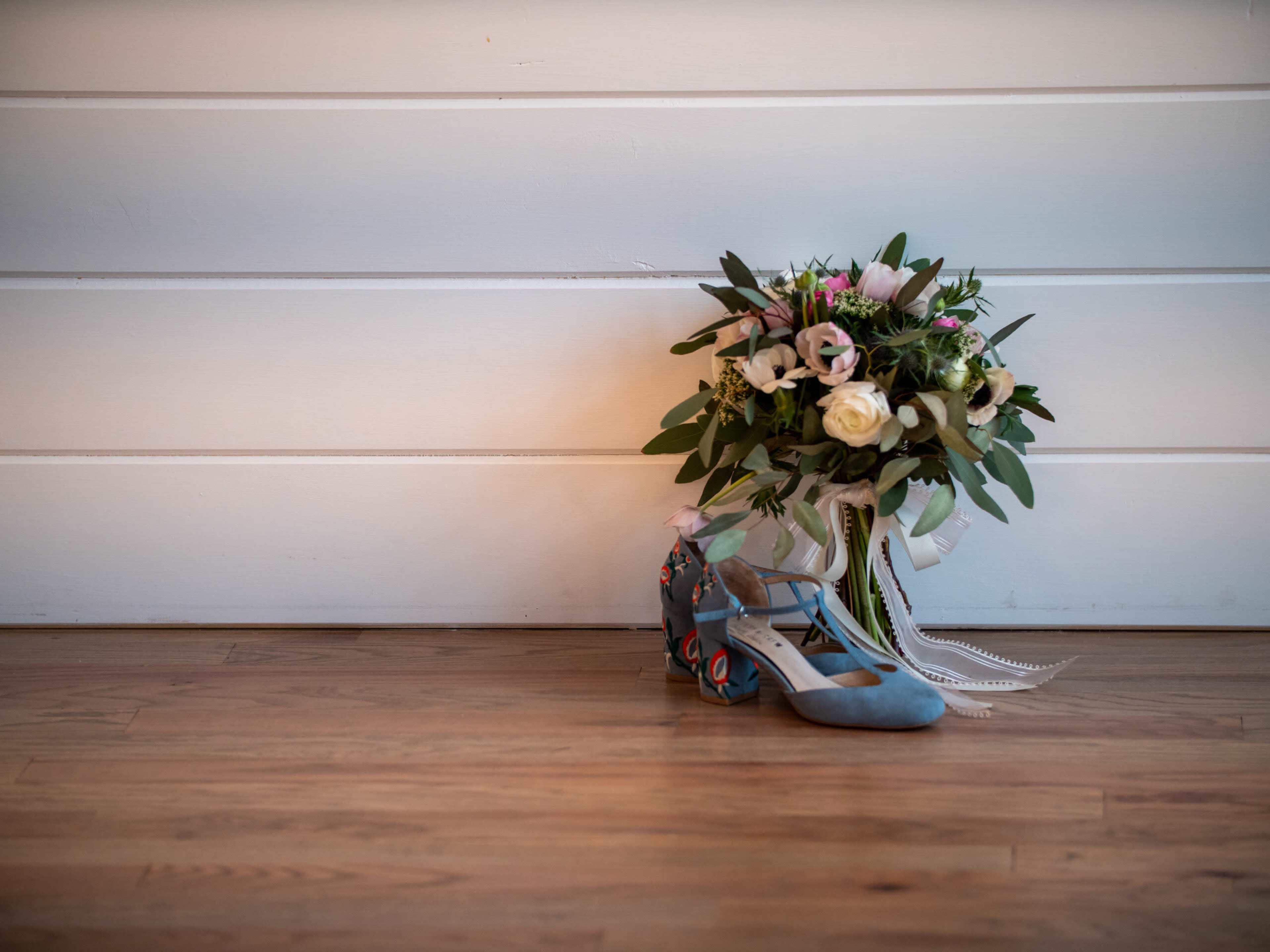 A pair of blue shoes sits next to a floral bouquet against a white wooden backdrop.