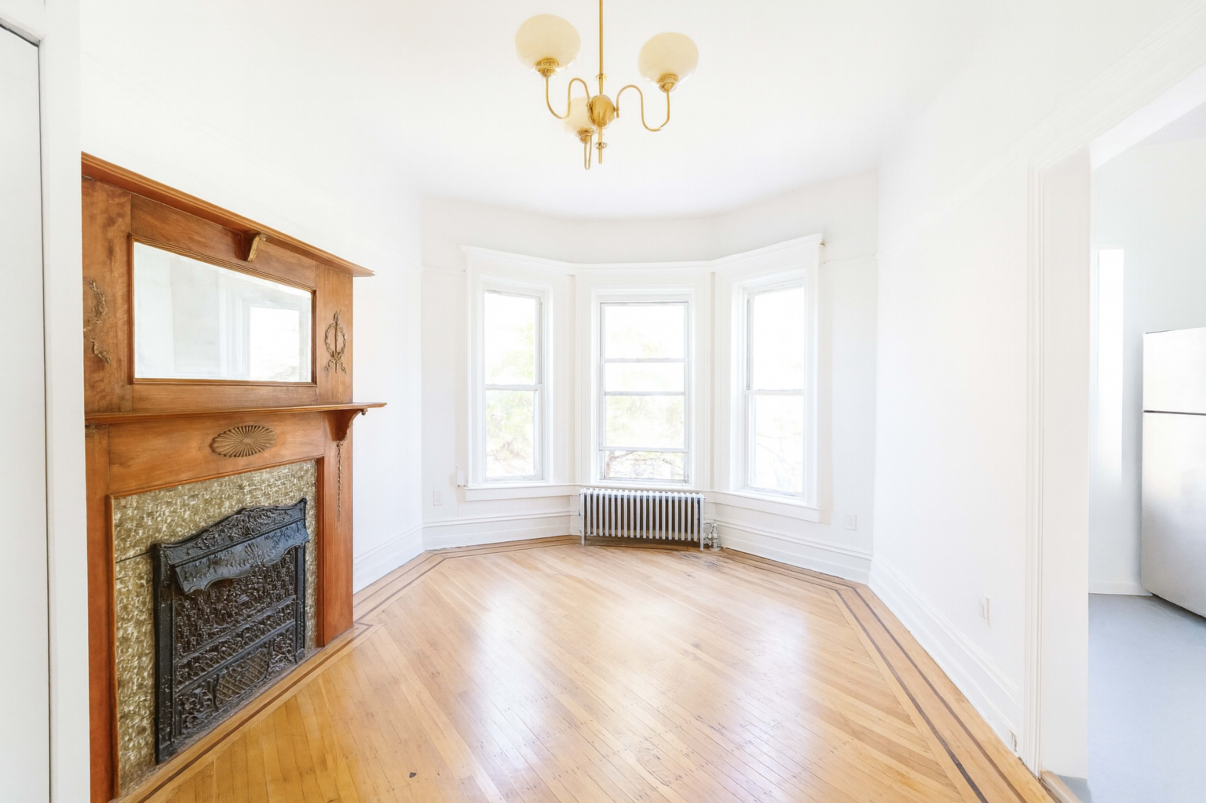 The image shows a bright, empty room with large windows, hardwood floors, and a decorative fireplace.