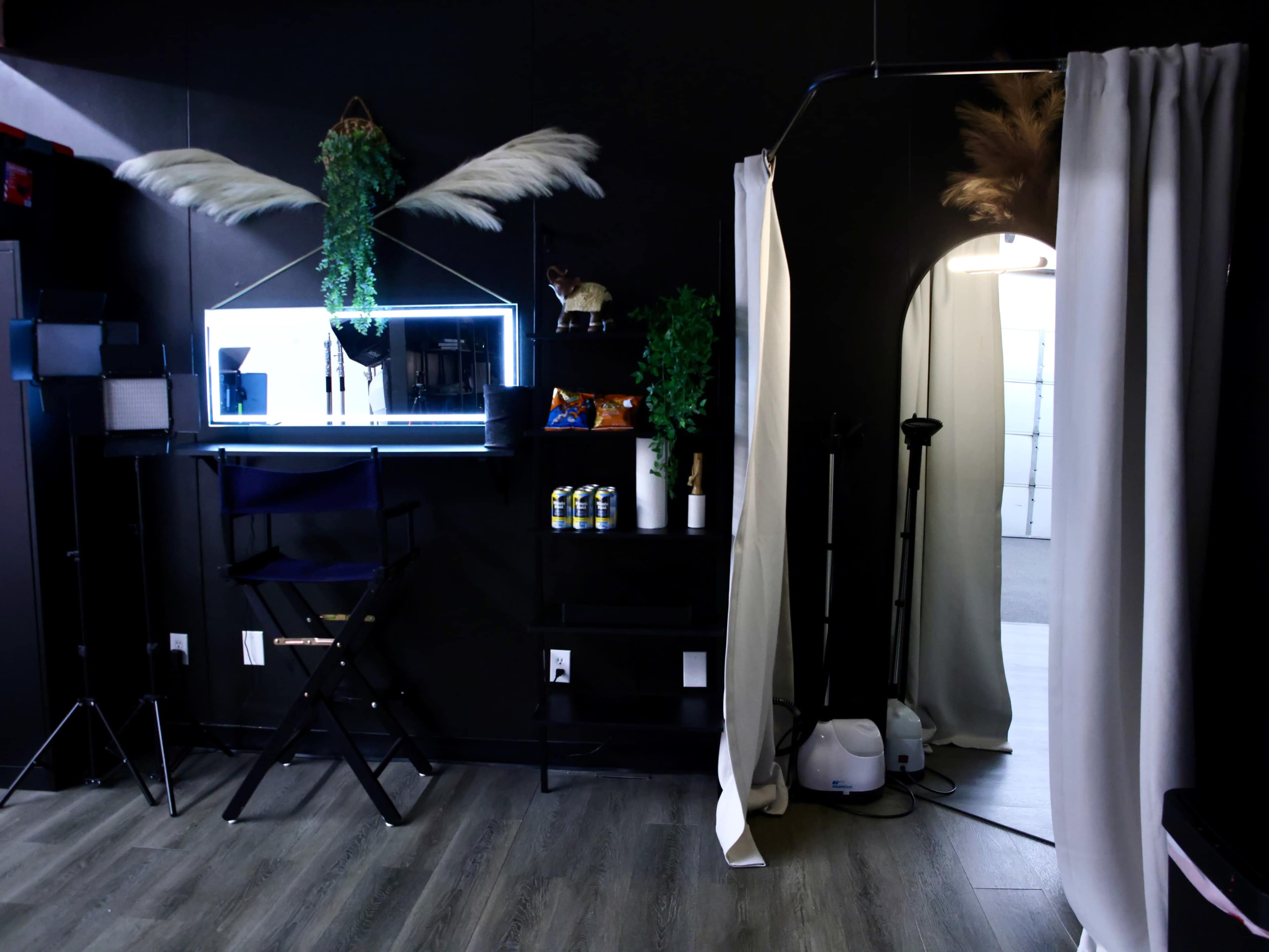 The image shows a dimly lit studio space with a black backdrop, a mirror with LED lights, a chair, shelves with snacks, and a doorway draped with a curtain.