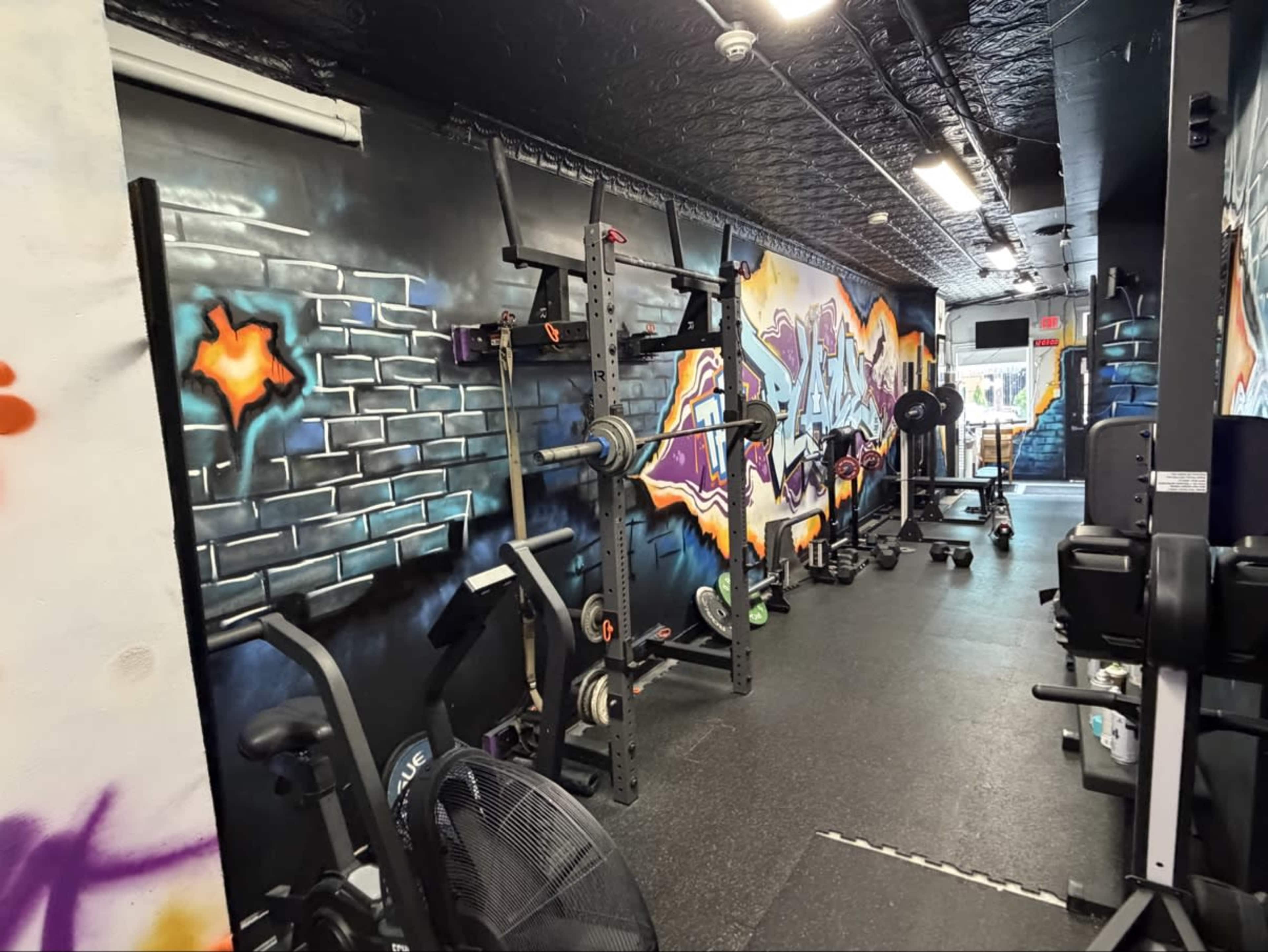 The image shows a gym with exercise equipment aligned against a graffiti-decorated wall.