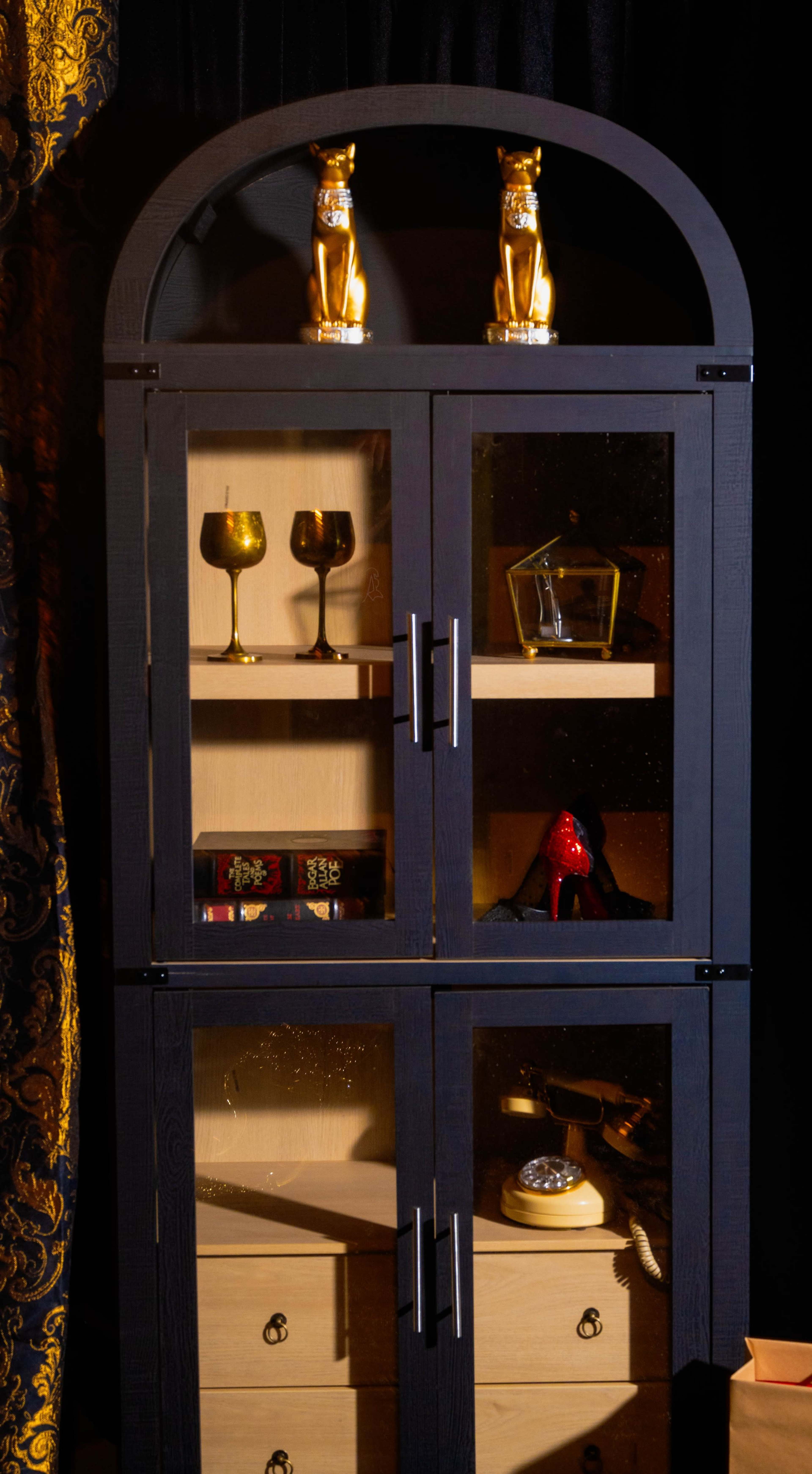 The image shows a two-tiered display cabinet featuring glass doors containing decorative items, including golden figurines, wine glasses, and a vintage telephone.