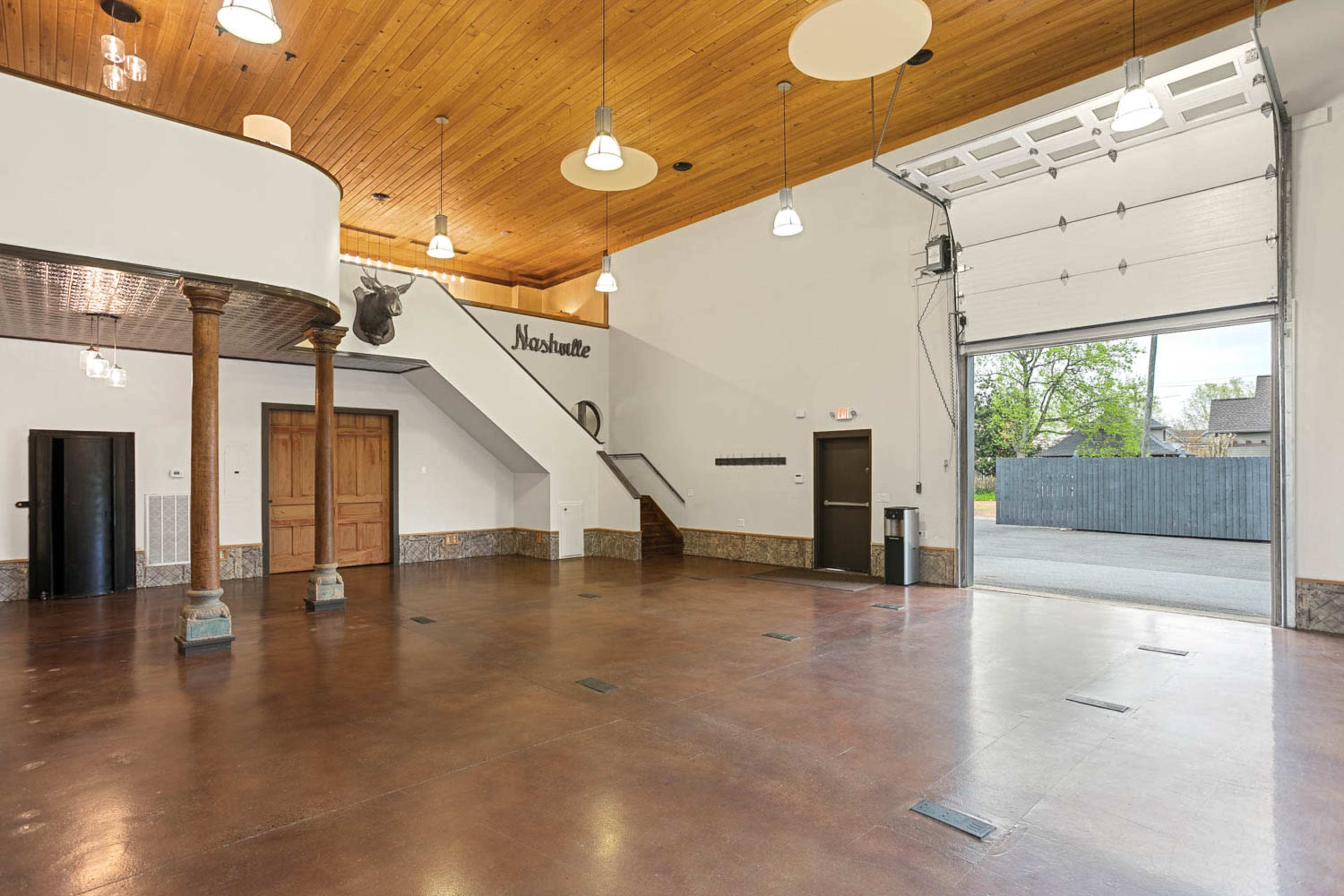 The interior of a spacious, modern building features a wooden ceiling, polished floor, and a staircase leading to an upper level with a large garage door opening to the outside.