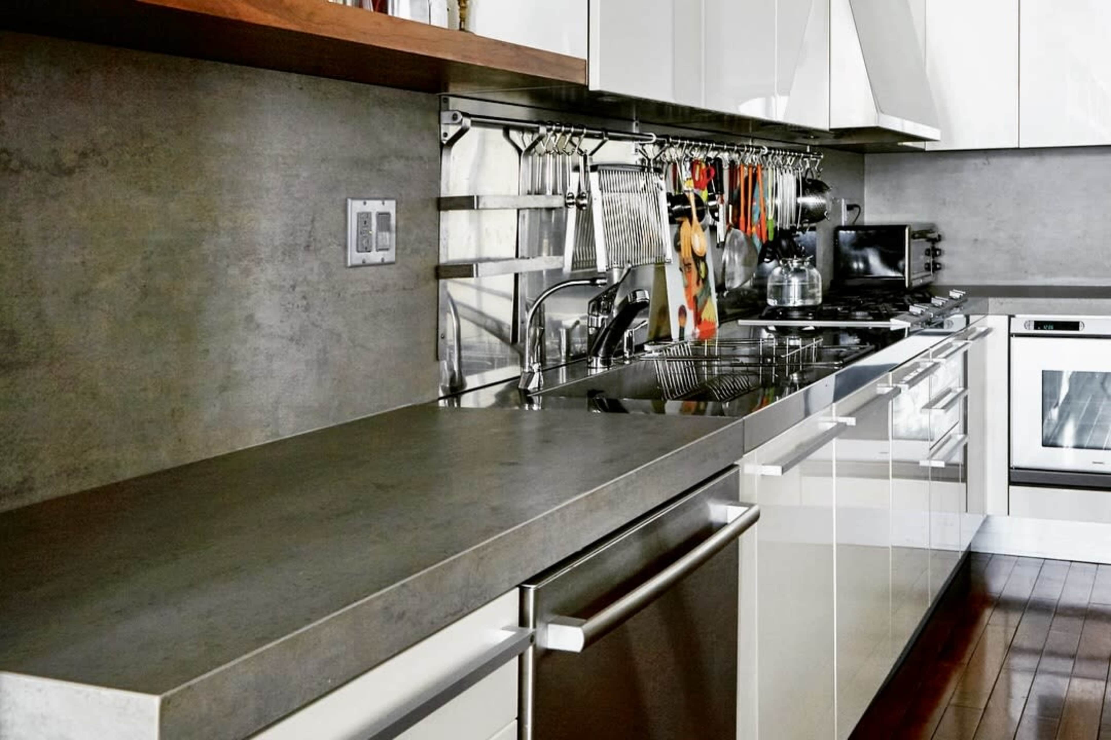 The image shows a modern kitchen with a concrete countertop, stainless steel appliances, and utensils hanging on a rack.