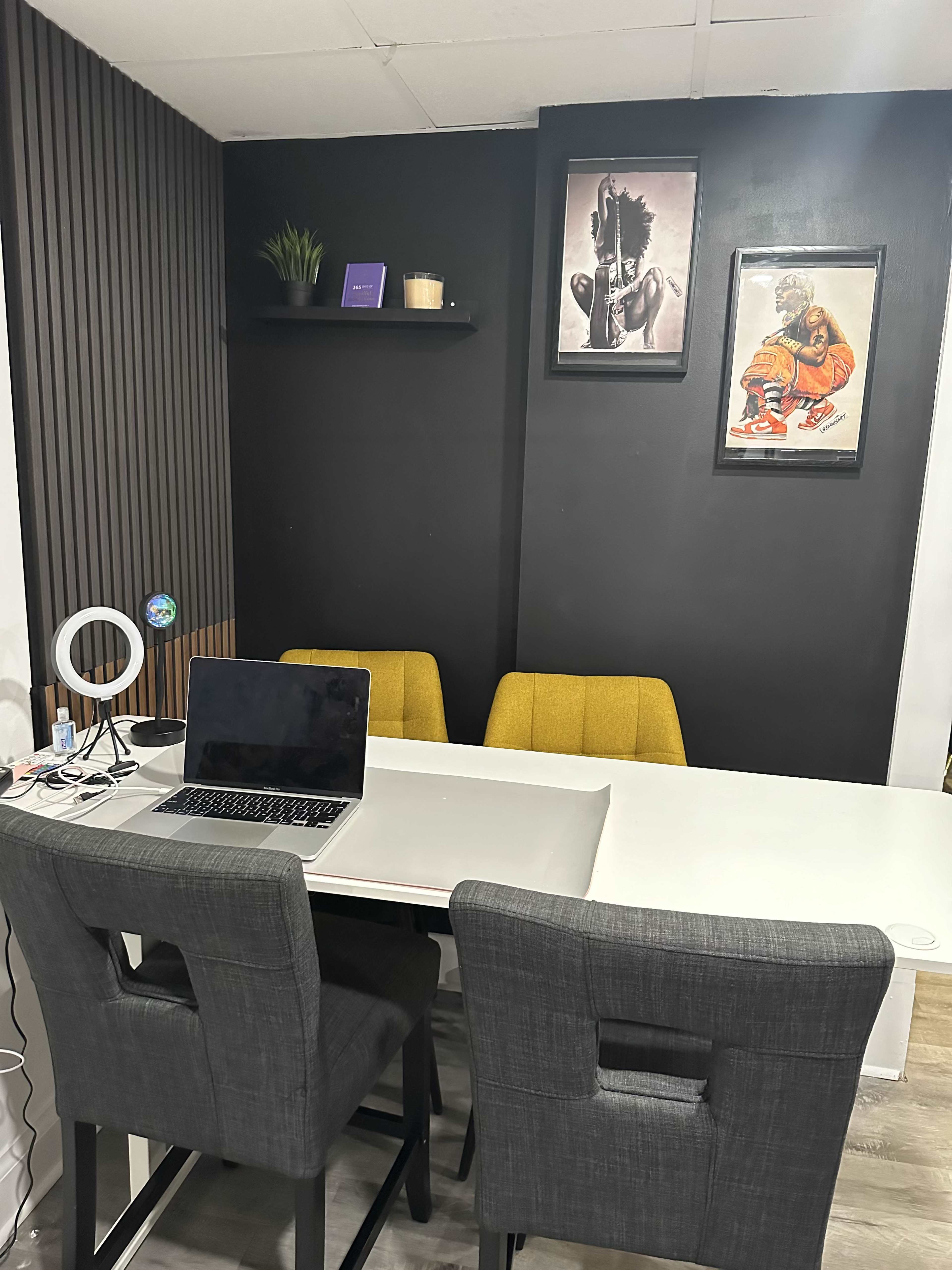 A modern office space with a white table, four chairs, a laptop, and two framed artworks on a black wall.