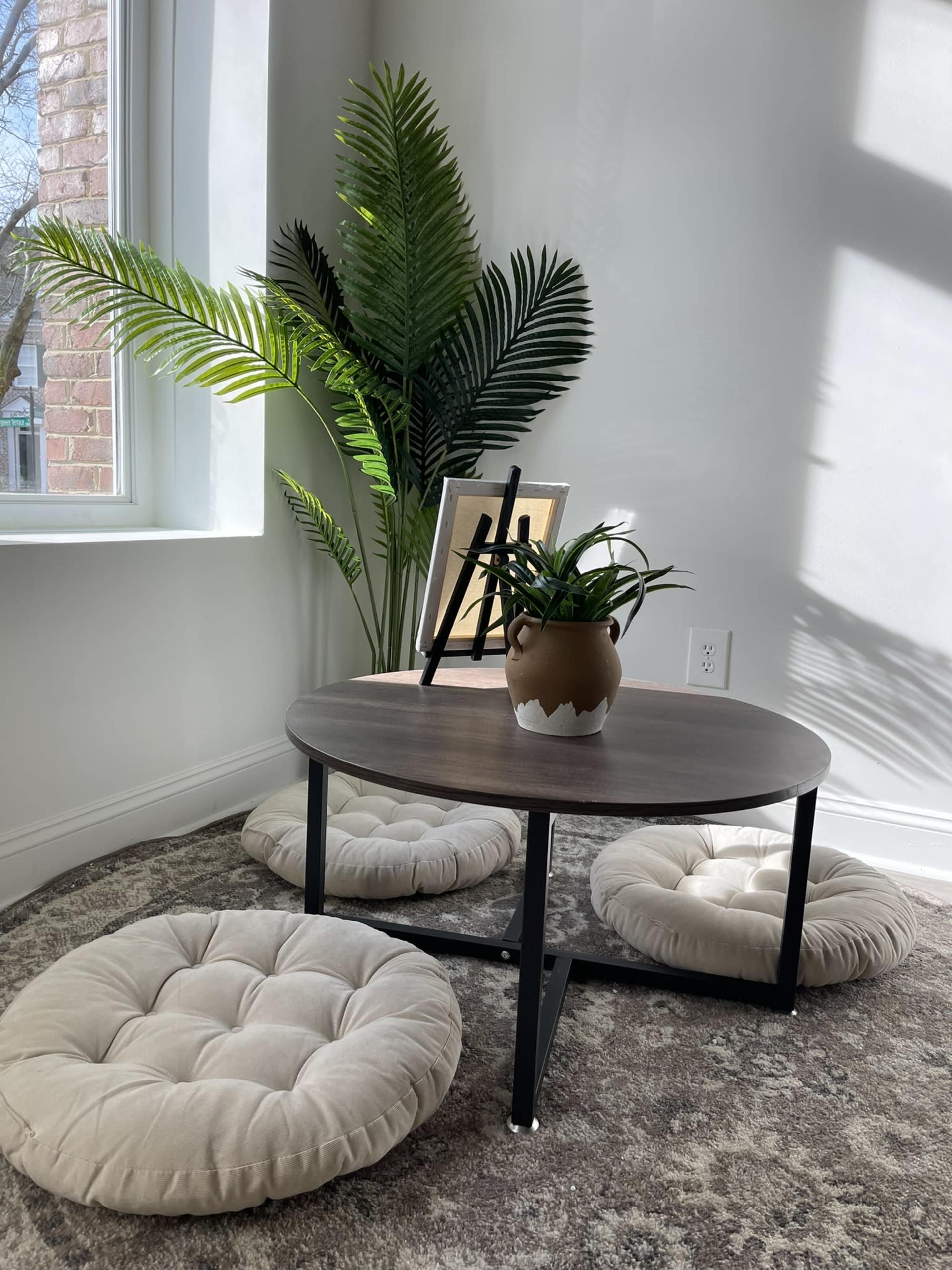 A small table is set in a well-lit corner with three round, cushioned floor seats, a potted plant, and a picture frame.