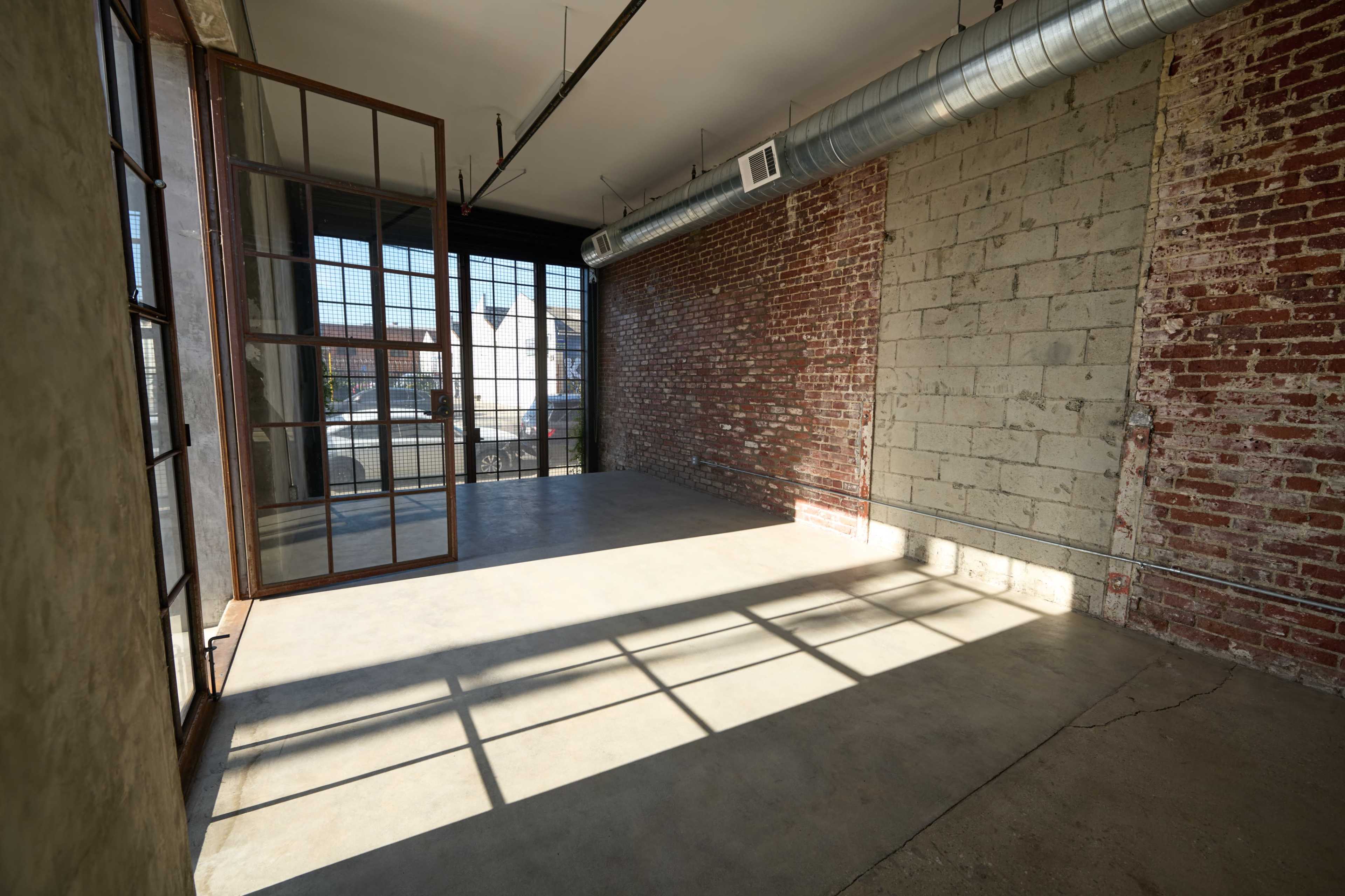 The image shows an empty industrial-style room with exposed brick walls, large windows, and a concrete floor.