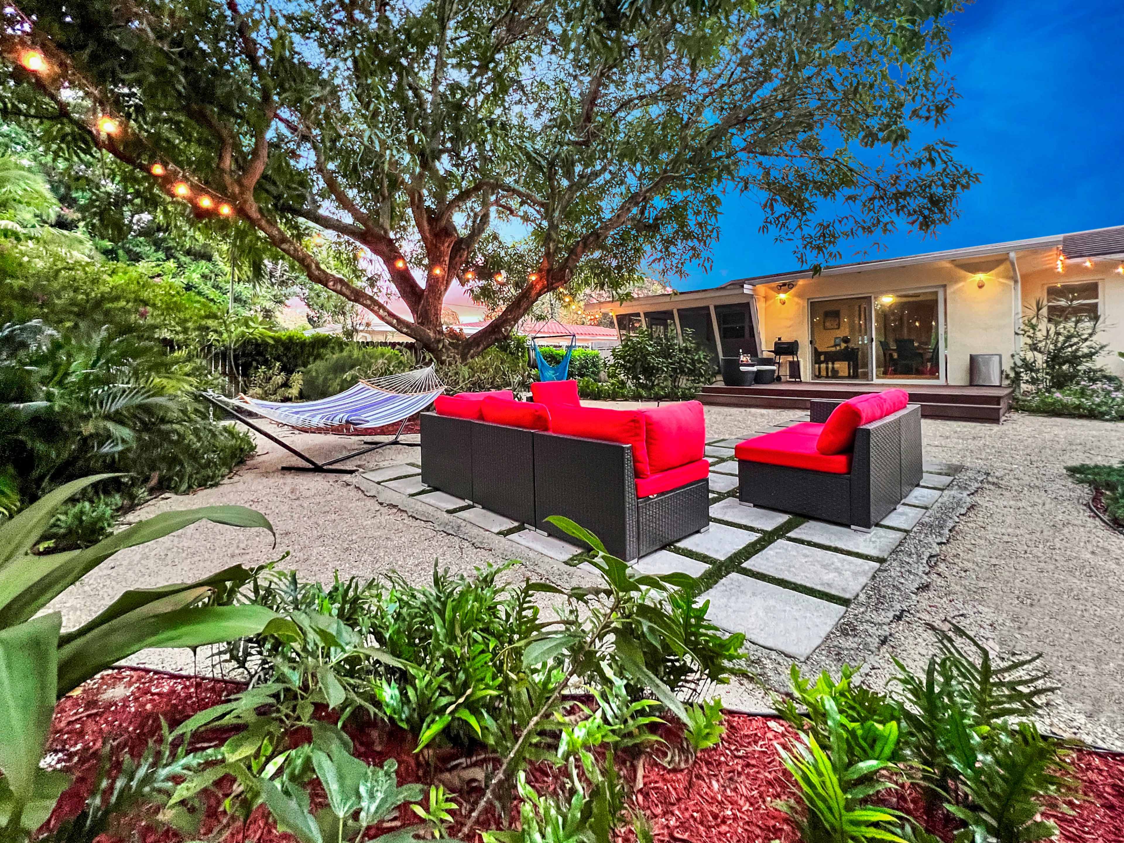 A cozy outdoor seating area features red cushions on black wicker furniture arranged around a stone path, with a hammock and garden greenery nearby.