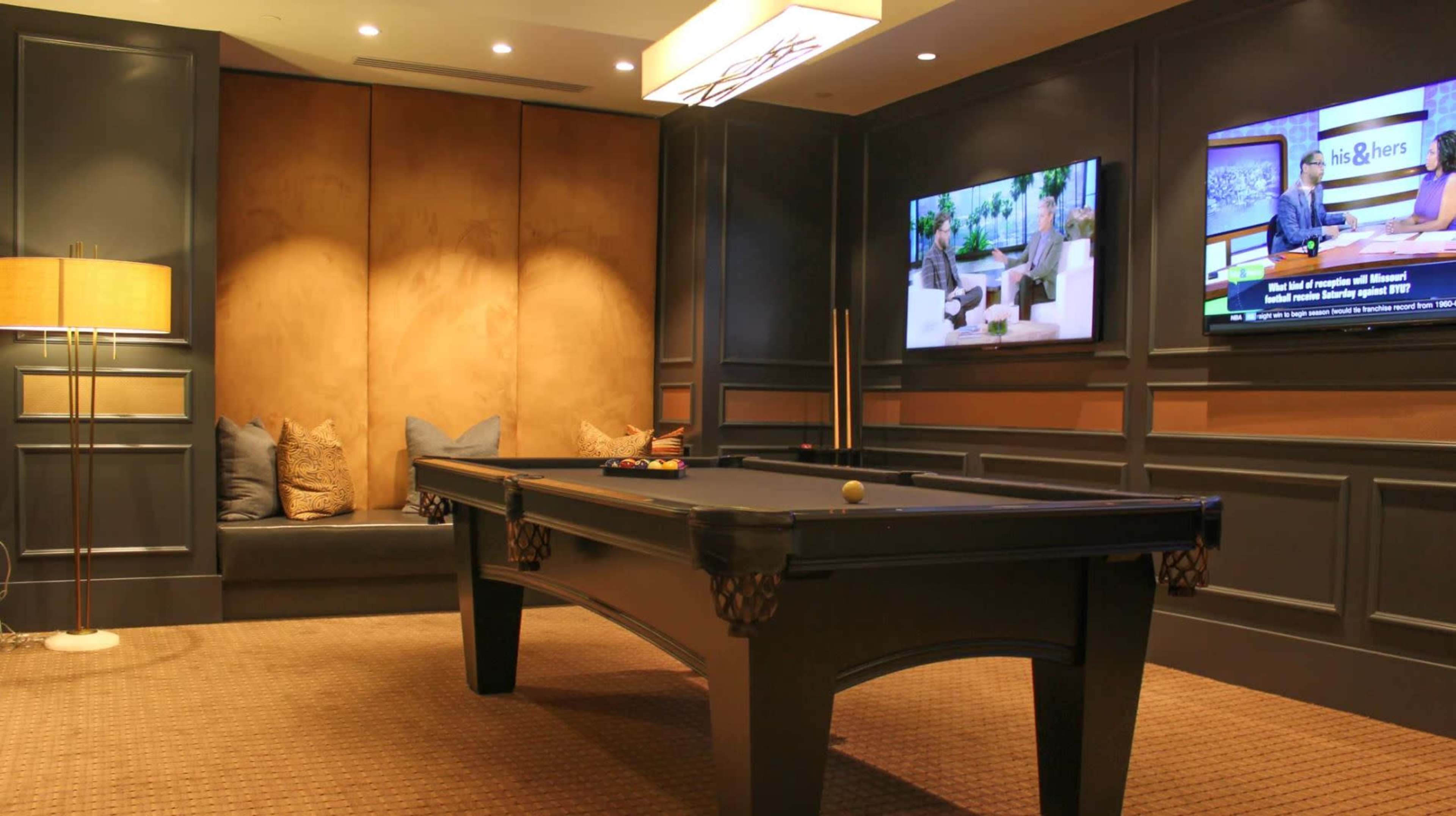 A modern game room features a pool table, a seating area with decorative pillows, and two wall-mounted televisions displaying programs.