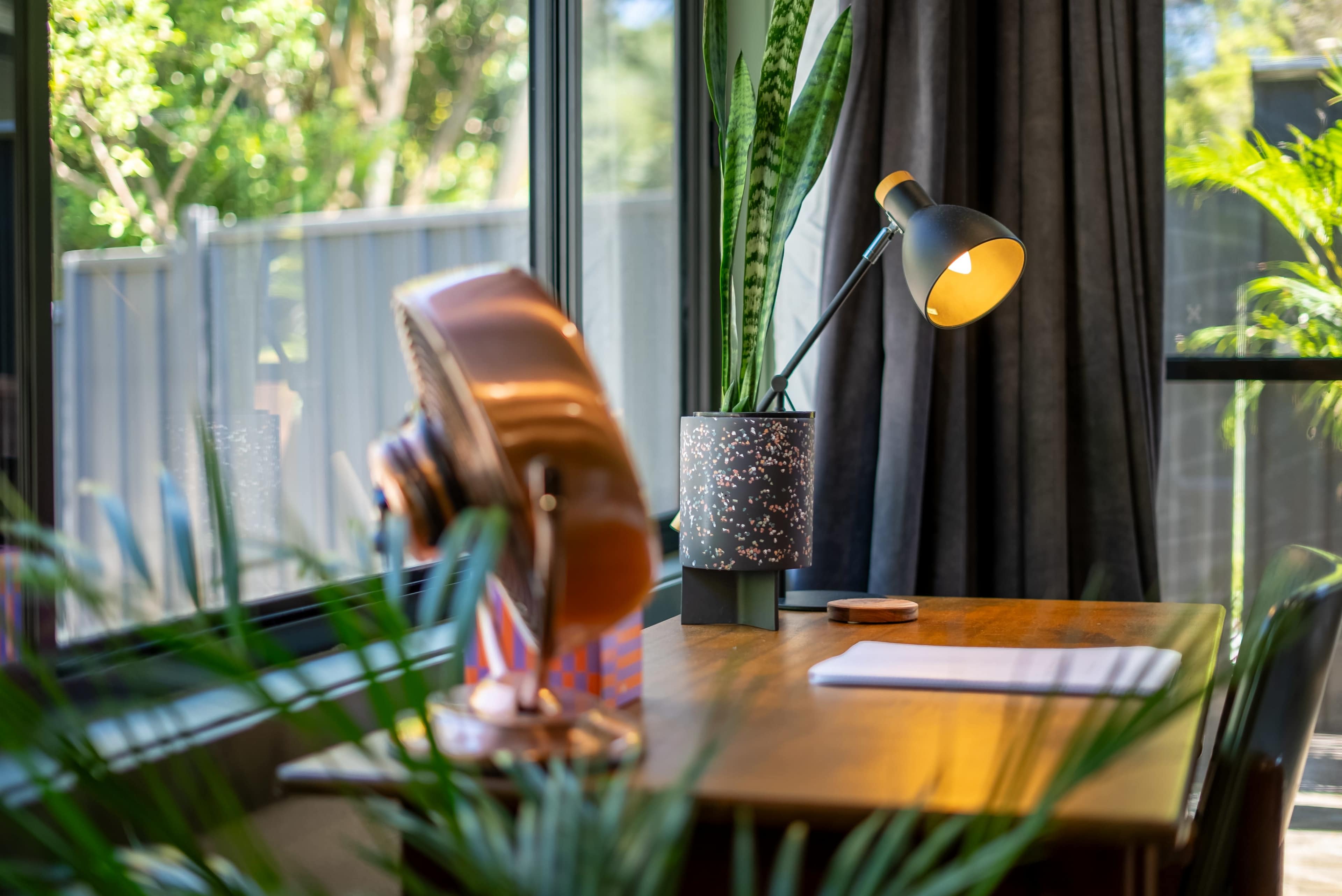 The image shows a wooden desk with a notebook and a lamp beside a potted plant, near a window with a view of greenery.