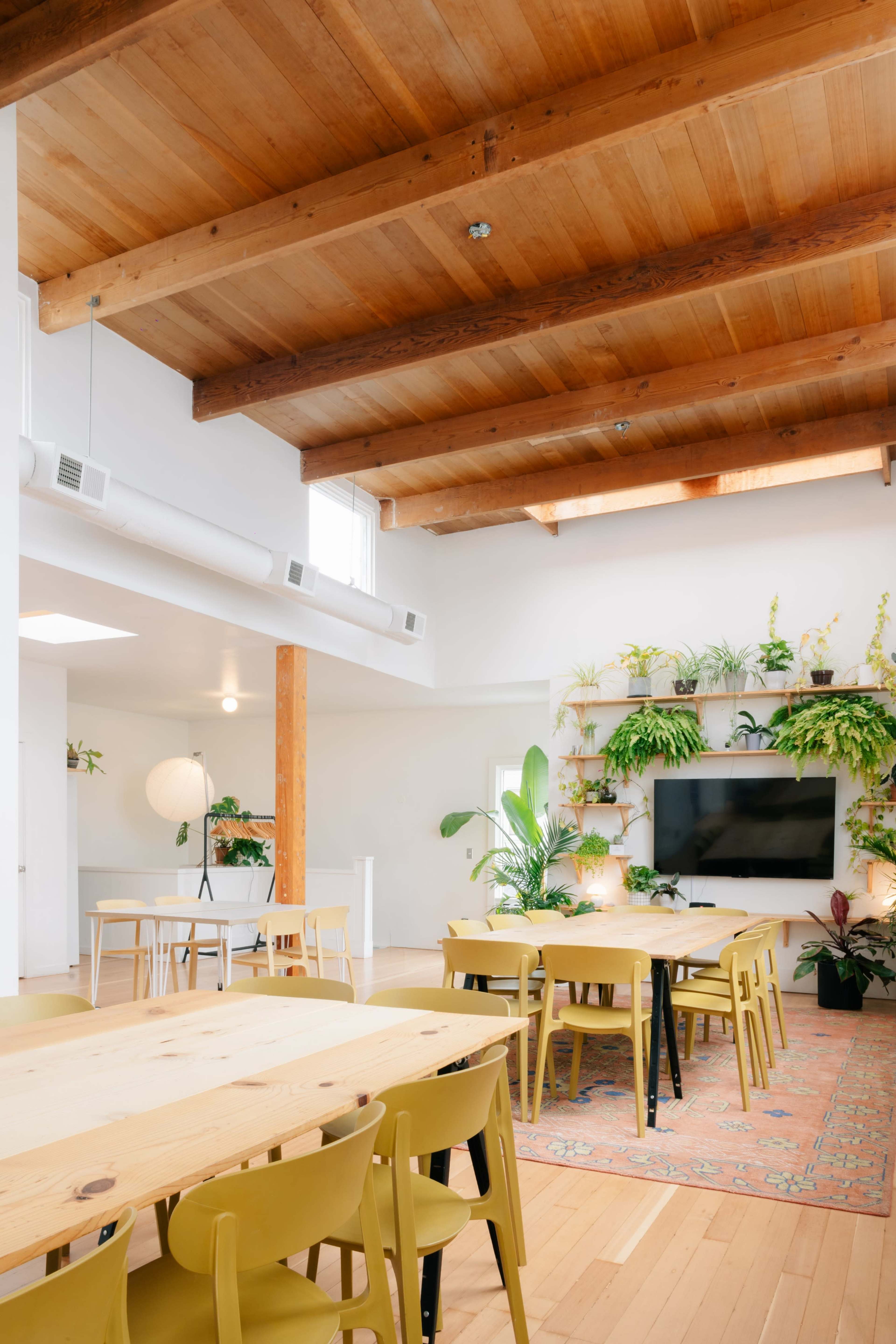 A spacious interior room features wooden beams, a large table with yellow chairs, and plants arranged around a television.
