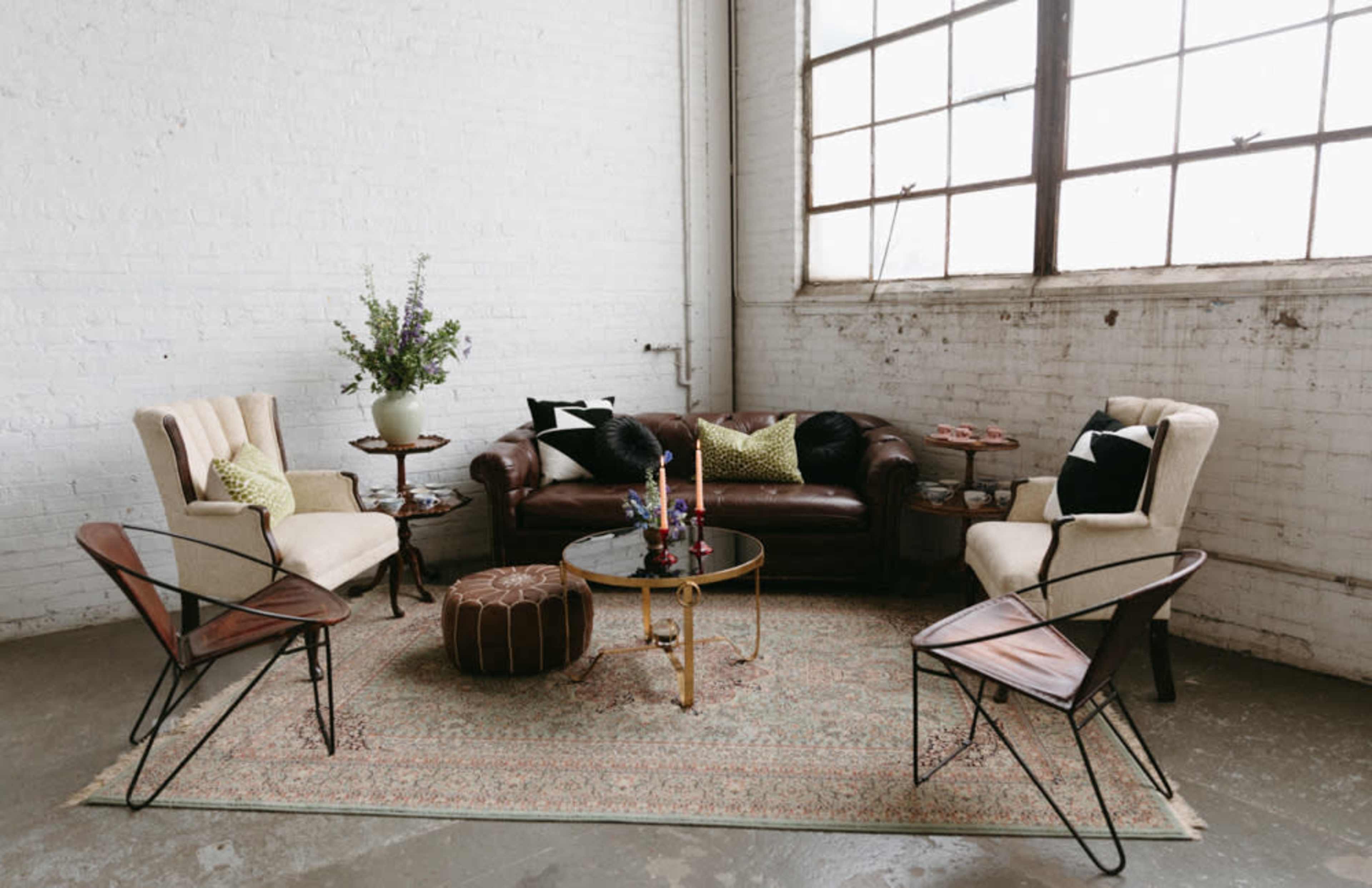 The image shows a cozy living room with a brown leather sofa, two upholstered armchairs, a round coffee table, and a patterned rug, all arranged near a large window.