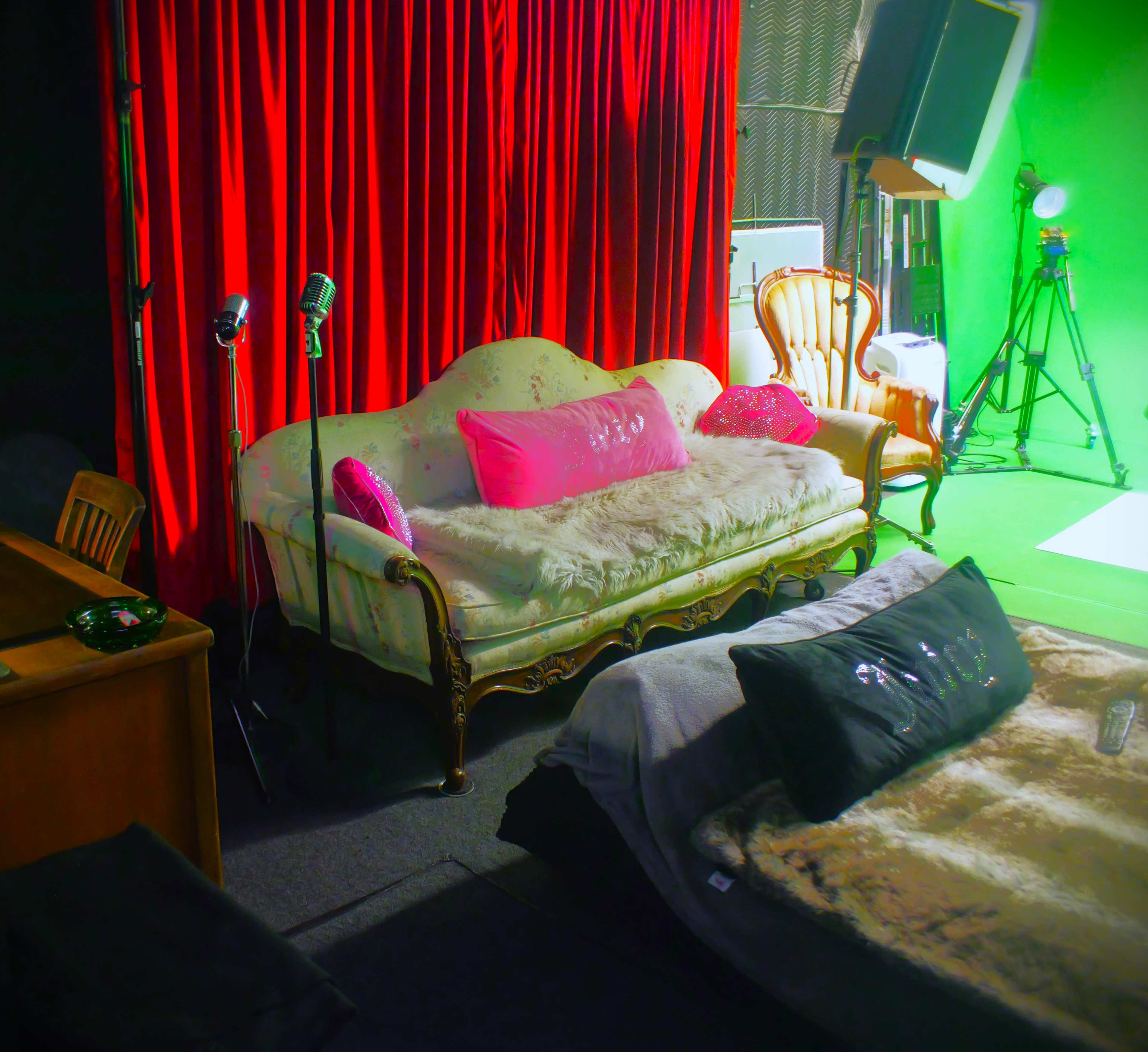The image shows a studio setting with a vintage sofa and chairs, draped in a red curtain, next to a green screen.