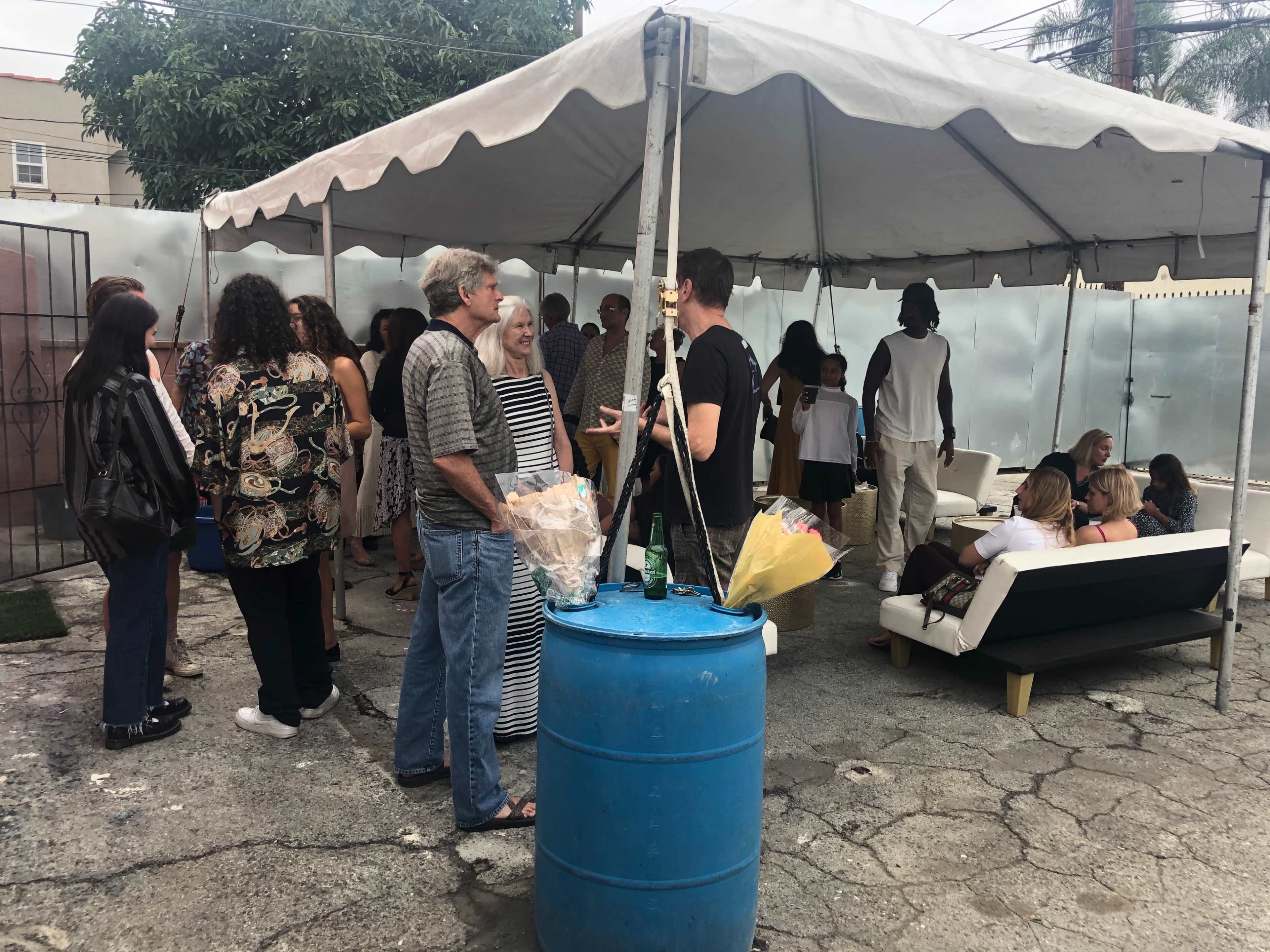A group of people is gathered under a tent in an outdoor space, with some standing near a blue barrel and others seated on furniture.