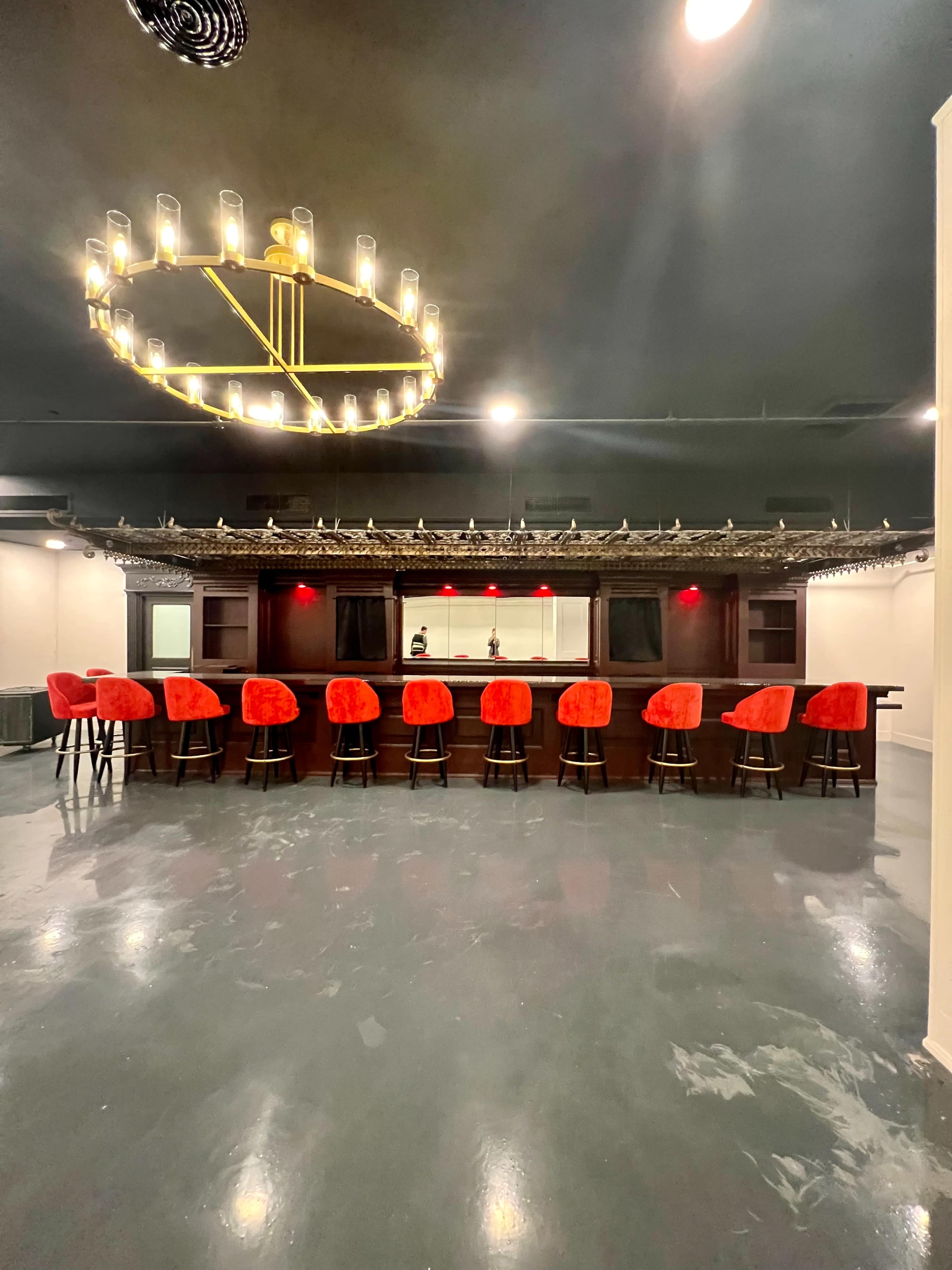 A dimly lit bar area with a circular chandelier above and ten red barstools lined up in front of a wooden bar.
