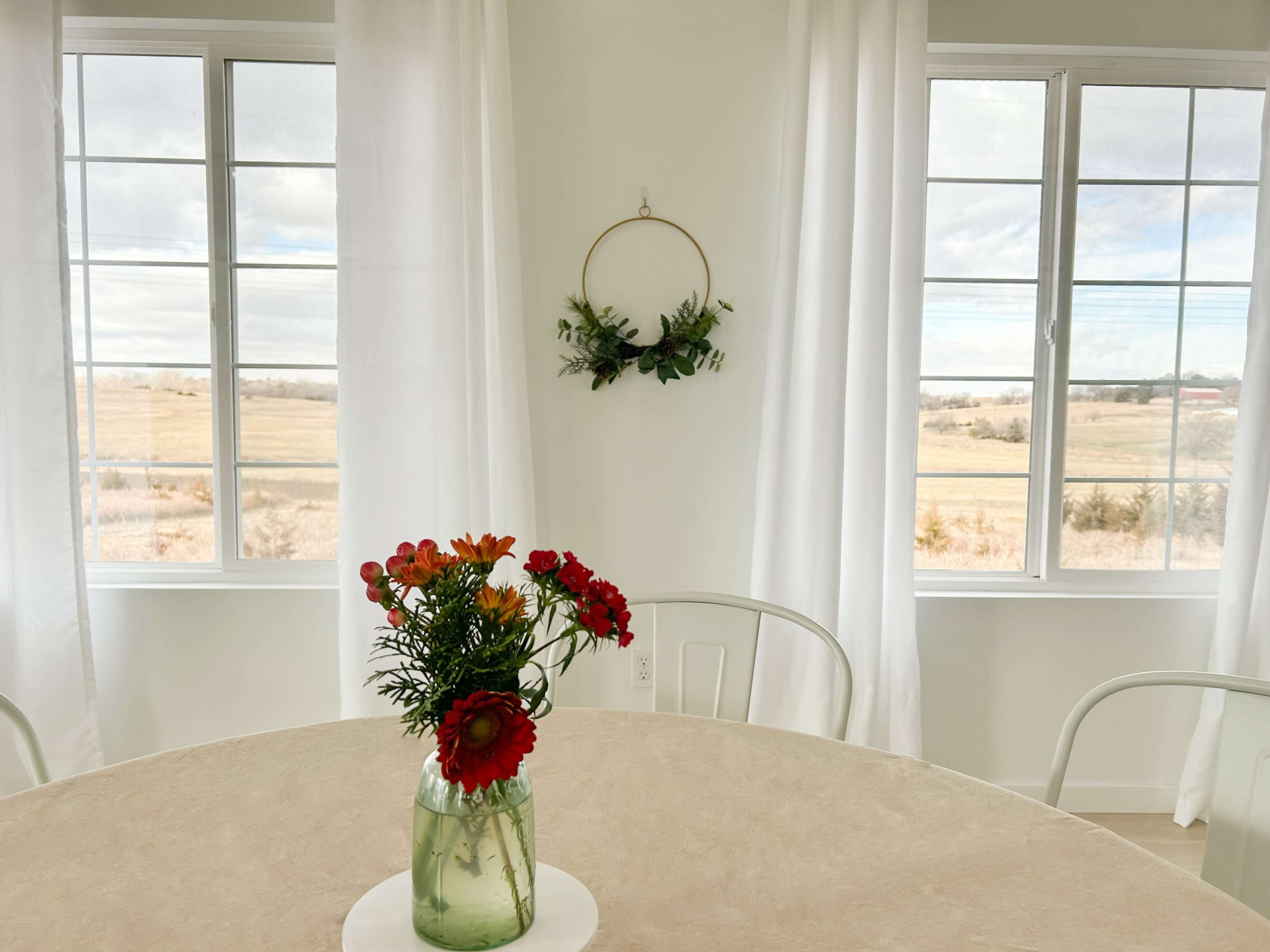 A simple table with a floral centerpiece is set against large windows that overlook a grassy field.
