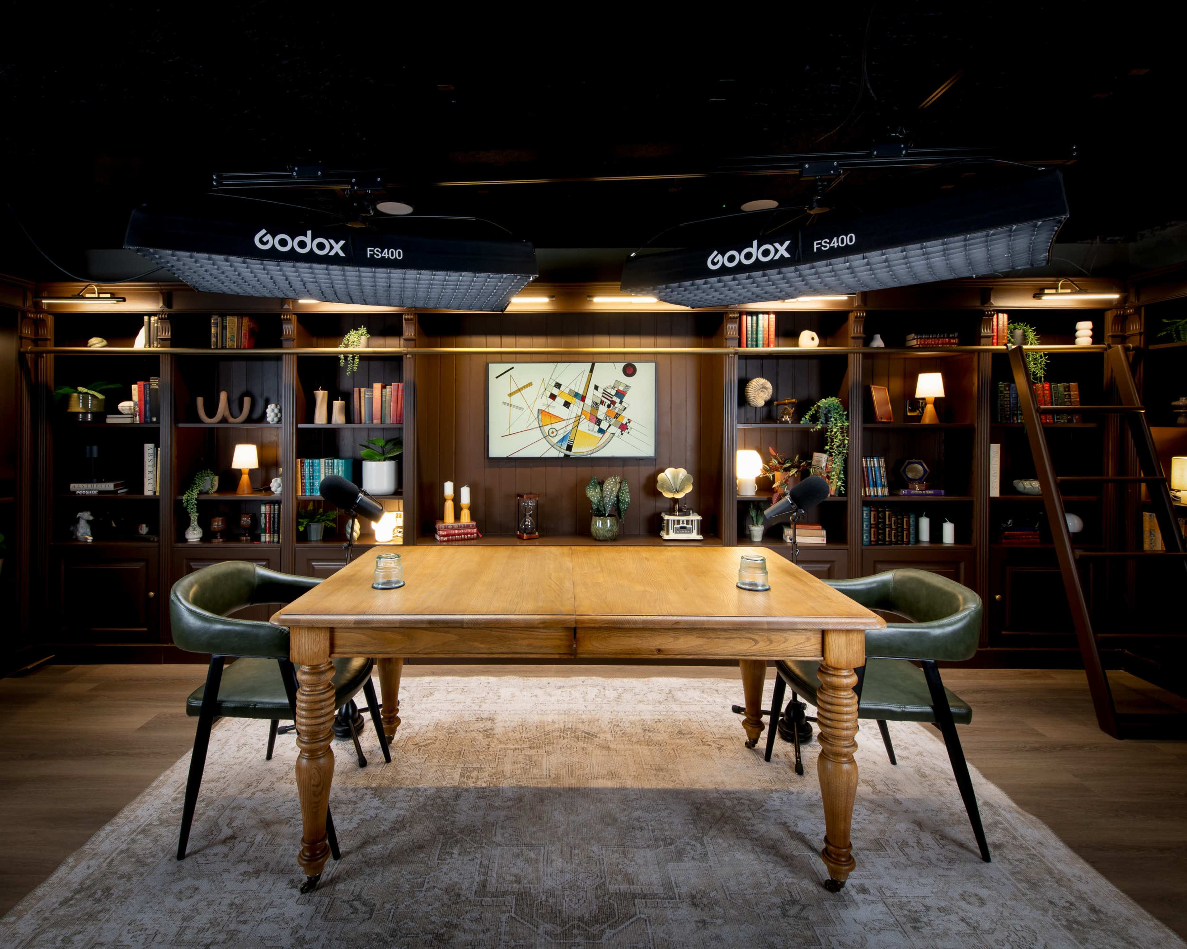 A well-lit room features a wooden table surrounded by two green chairs, with shelves lined with books and decorative items in the background.