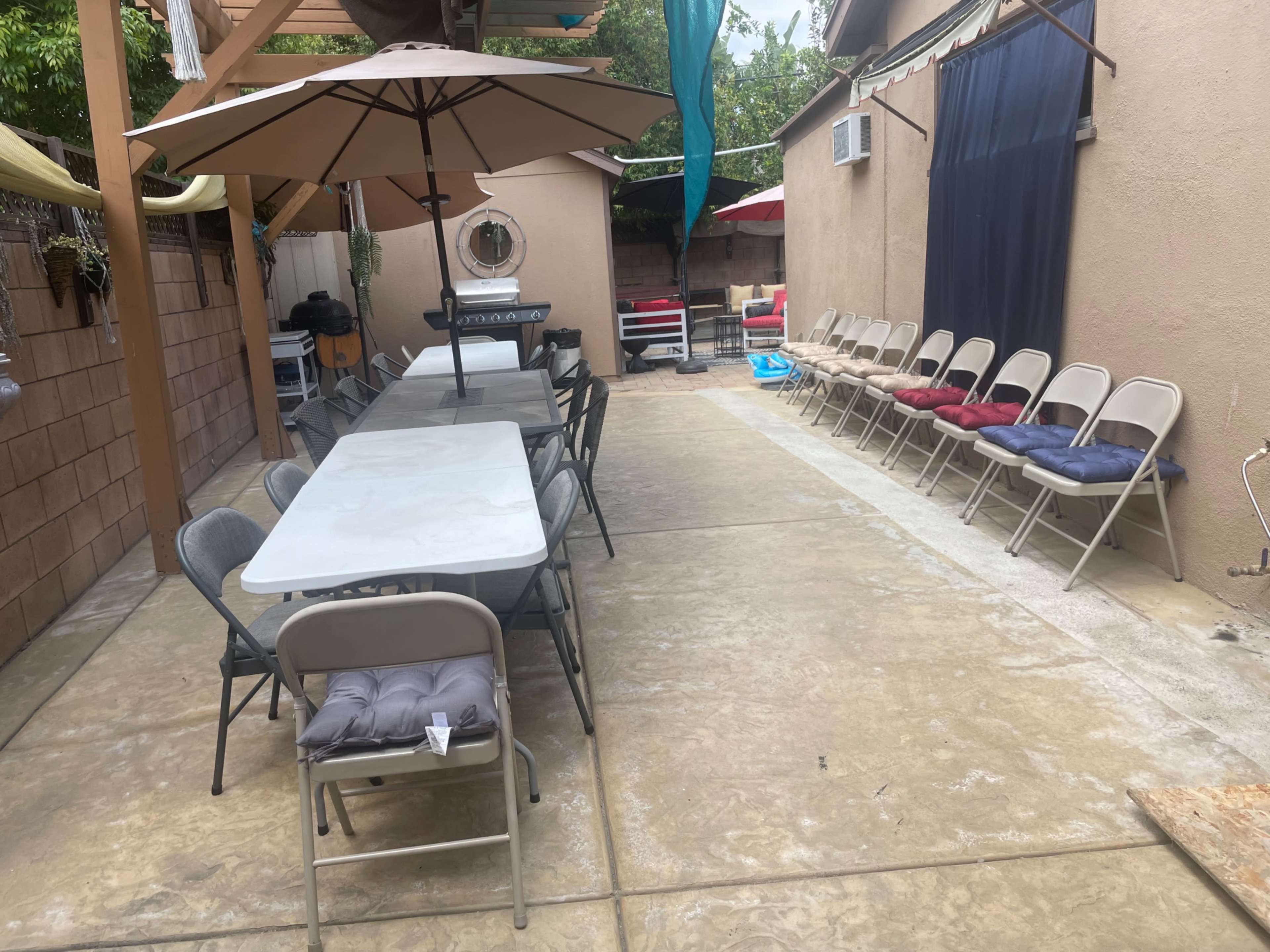 The image shows a patio area featuring a long table with chairs on one side and an umbrella overhead, with additional chairs arranged along the opposite wall.