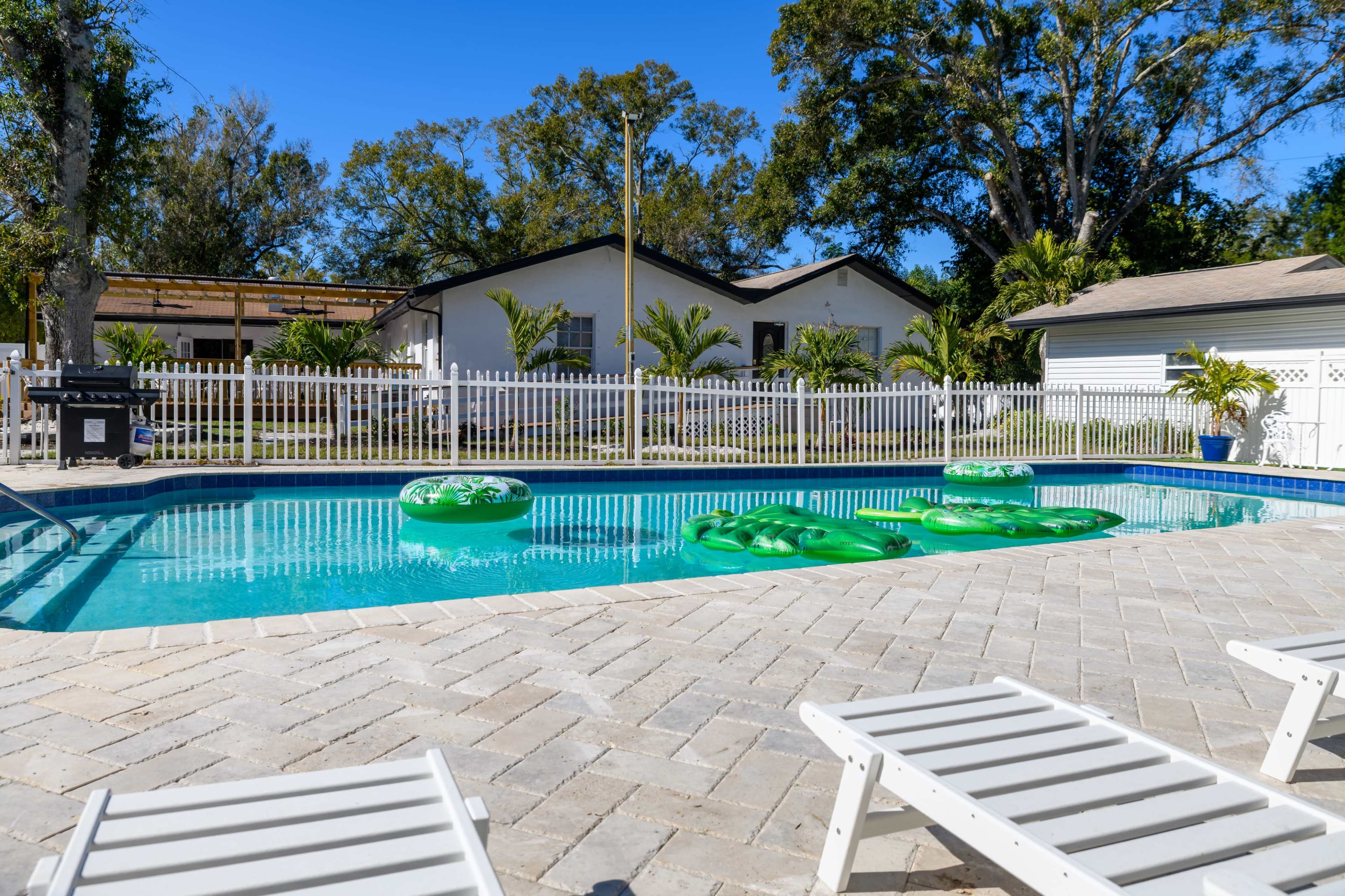 The image shows a swimming pool with green inflatable floats, surrounded by a fenced yard and several lounge chairs under a clear blue sky.