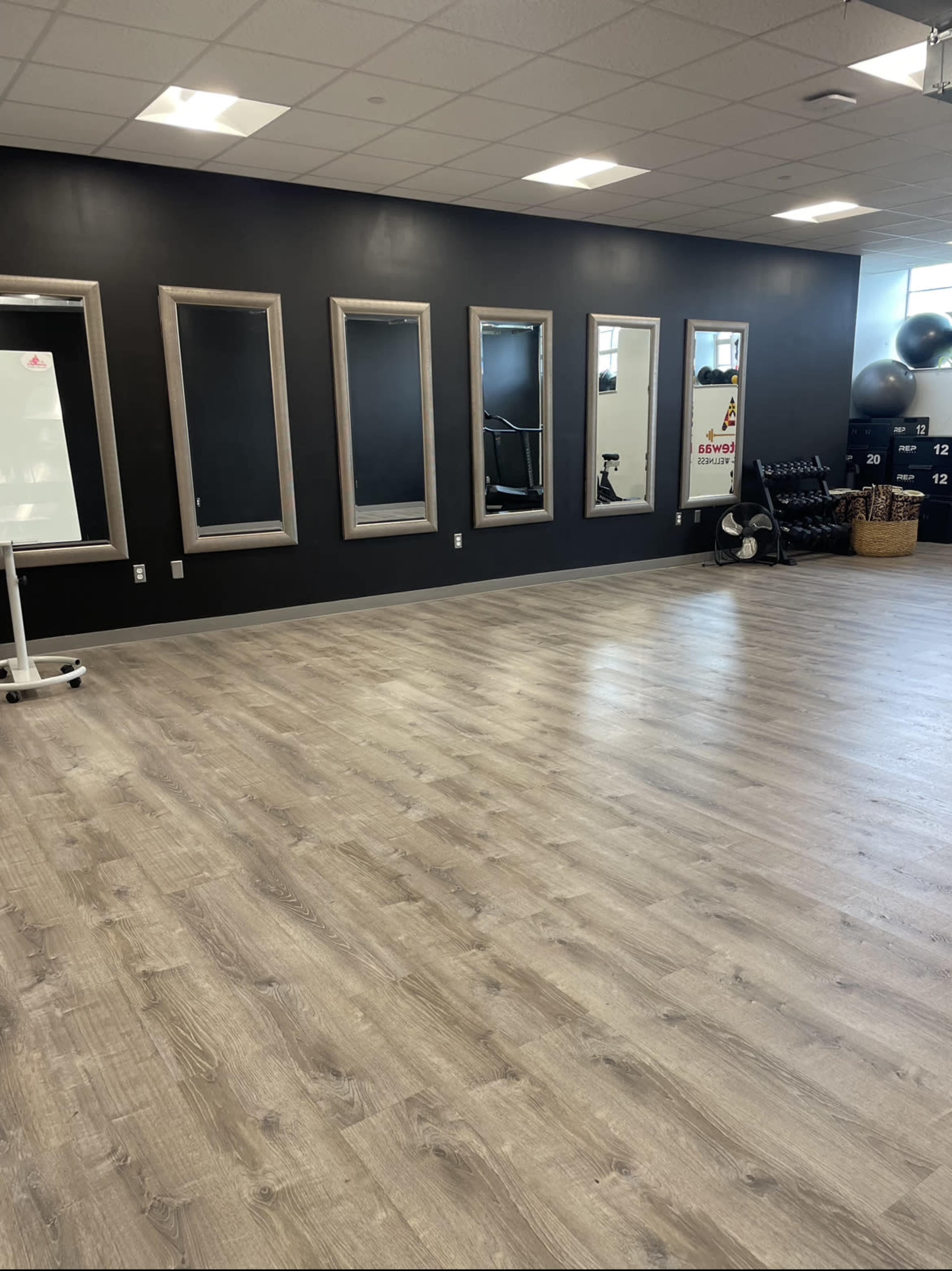 A fitness studio with wood-style flooring, black walls, and several mirrors on one side, along with exercise equipment and a whiteboard.