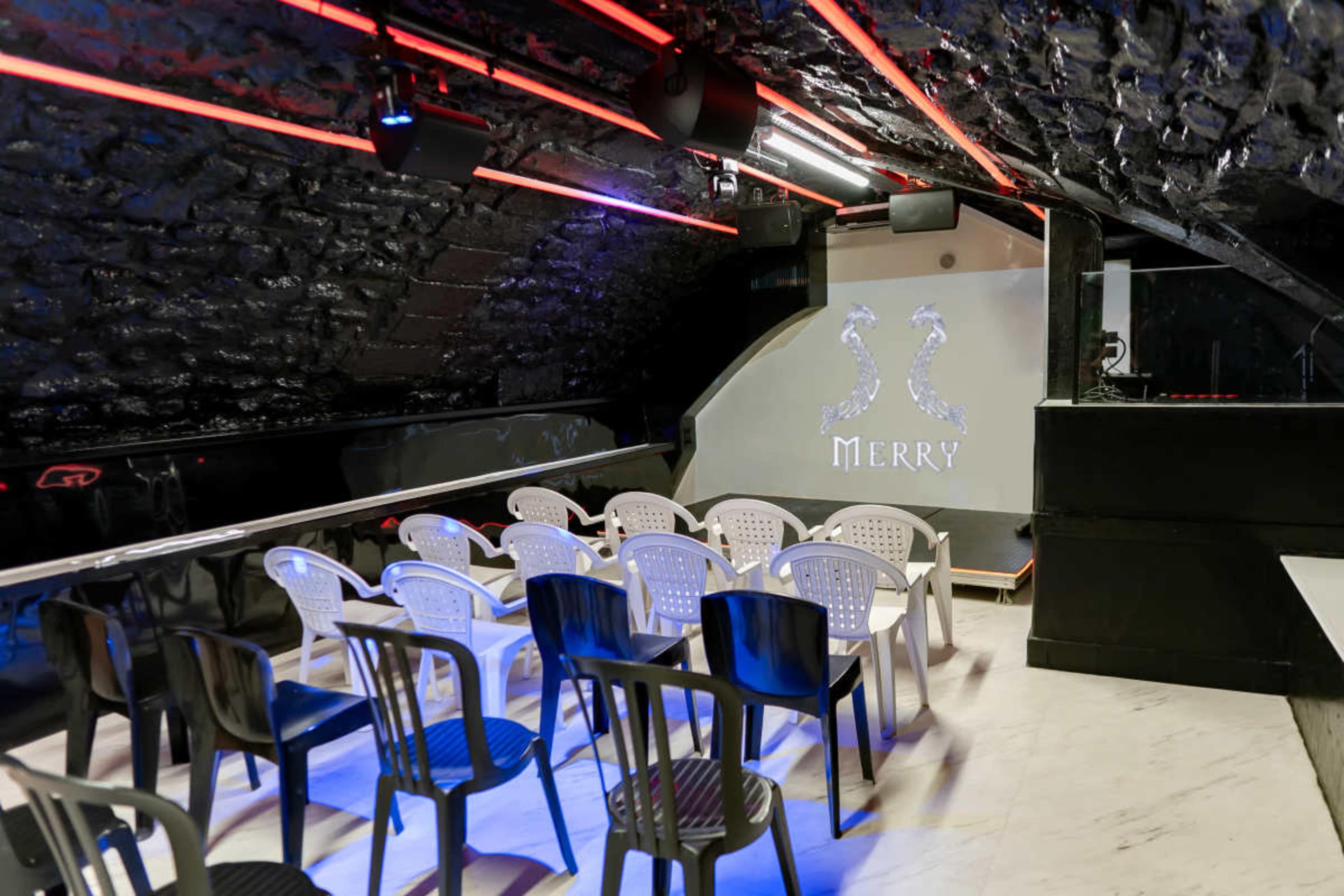 The image shows a small performance space with rows of plastic chairs under a dimly lit, vaulted ceiling, featuring a wall displaying the word "MERRY."