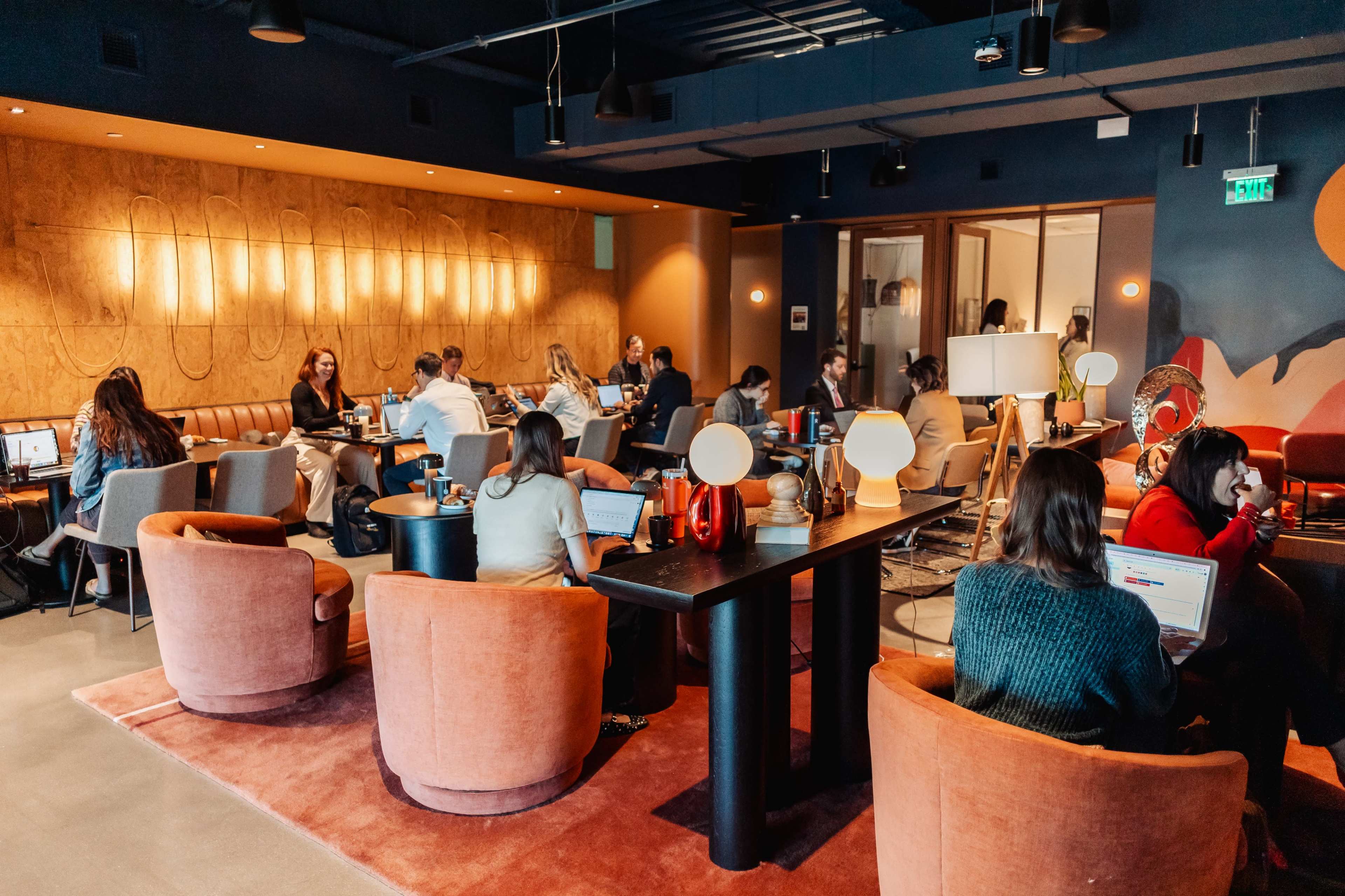 The image shows a busy coworking space with several people working on laptops at tables and comfortable seating areas.