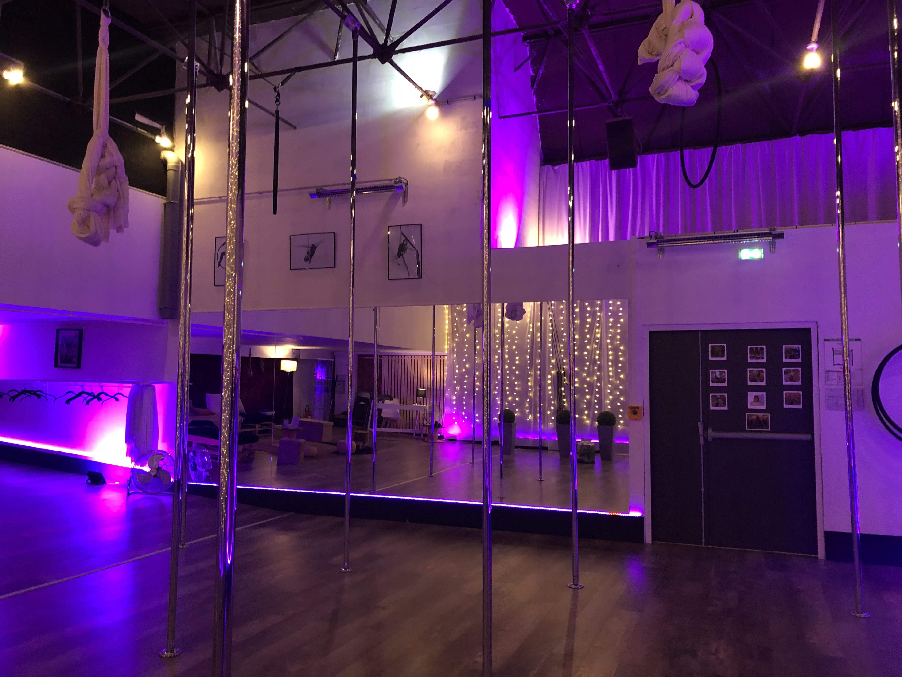 The image shows a spacious studio with several pole fixtures, mirrors, and ambient lighting, featuring purple hues and a sparkling backdrop.