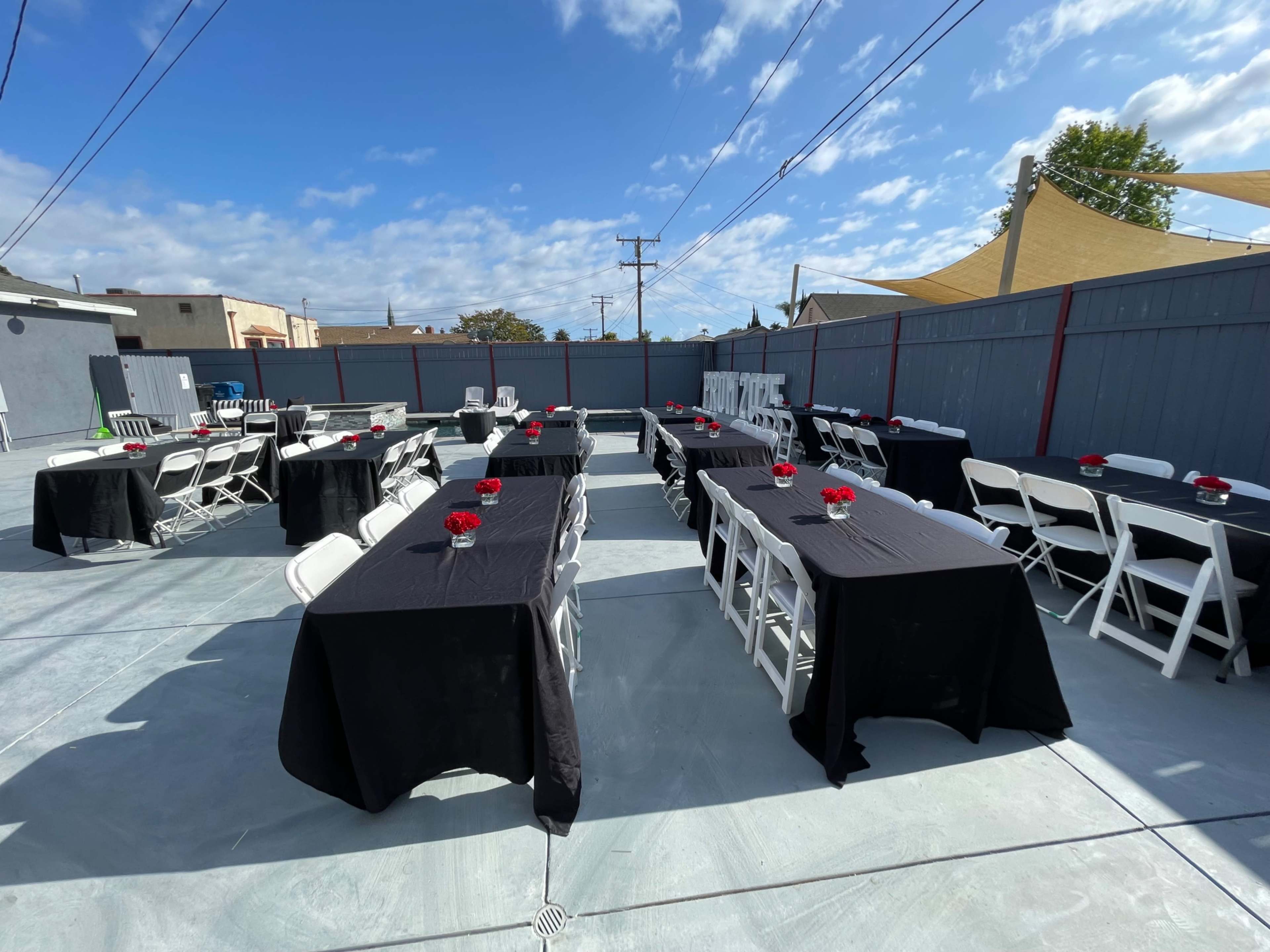 The image shows a rooftop outdoor event space set up with tables covered in black cloth, adorned with small red flower centerpieces, under a partly cloudy sky.