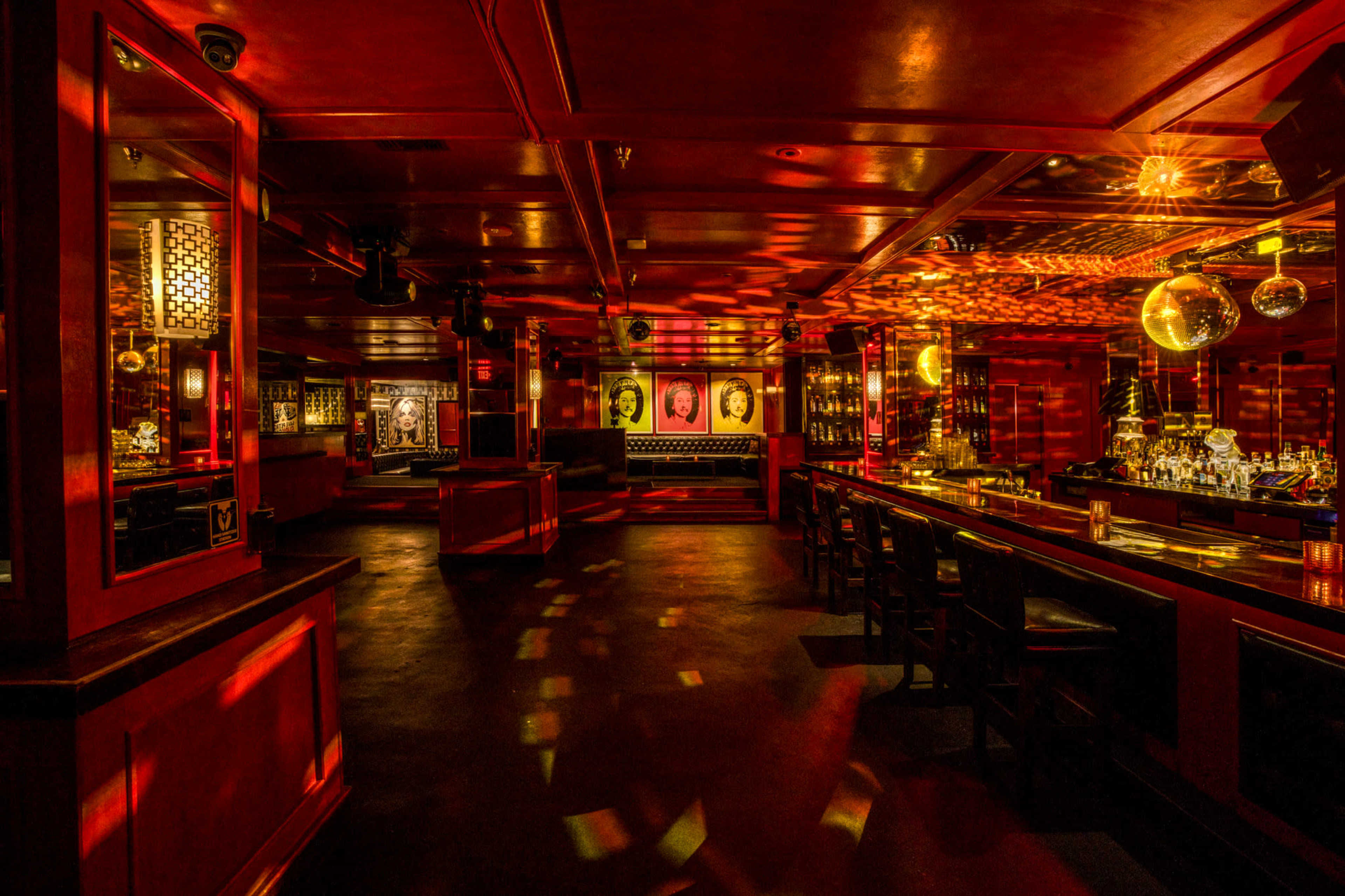 The image shows a dimly lit bar with red walls, disco balls, and an empty seating area, featuring illuminated artwork on the walls.