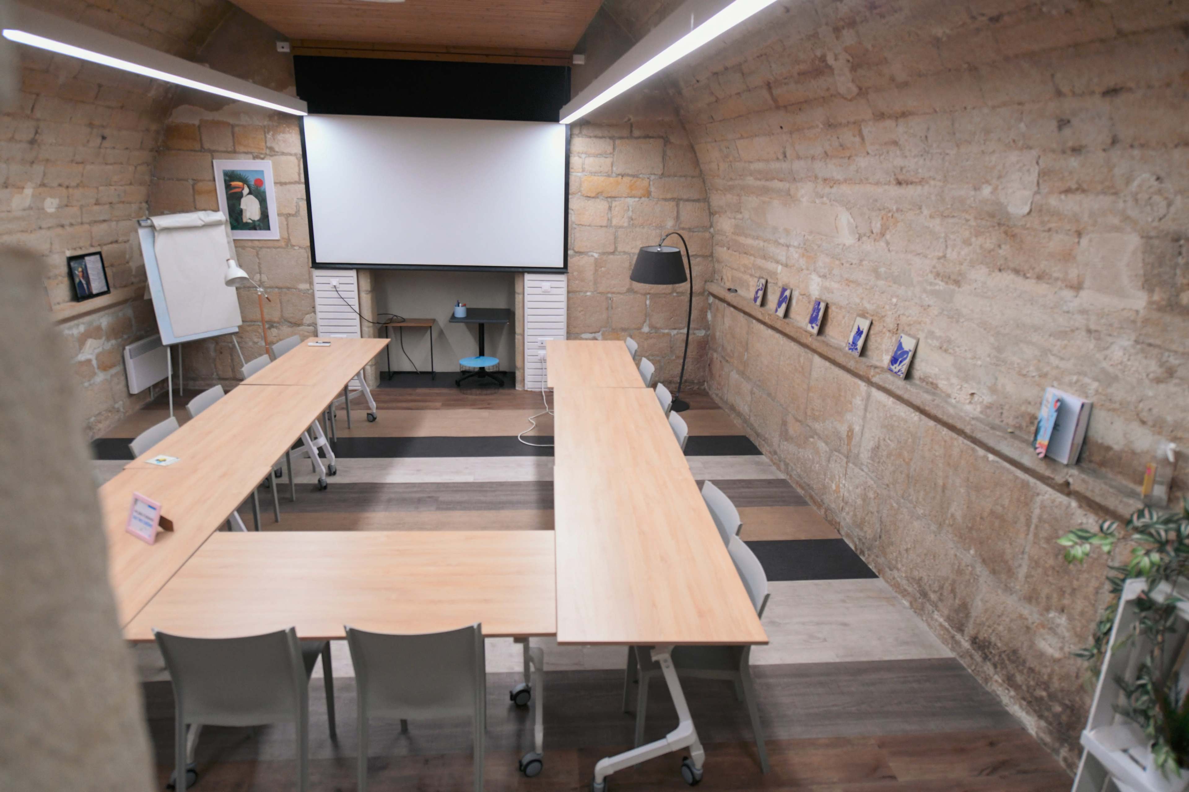 An empty meeting room with wooden tables arranged in a U shape, a large screen at the front, and stone walls.