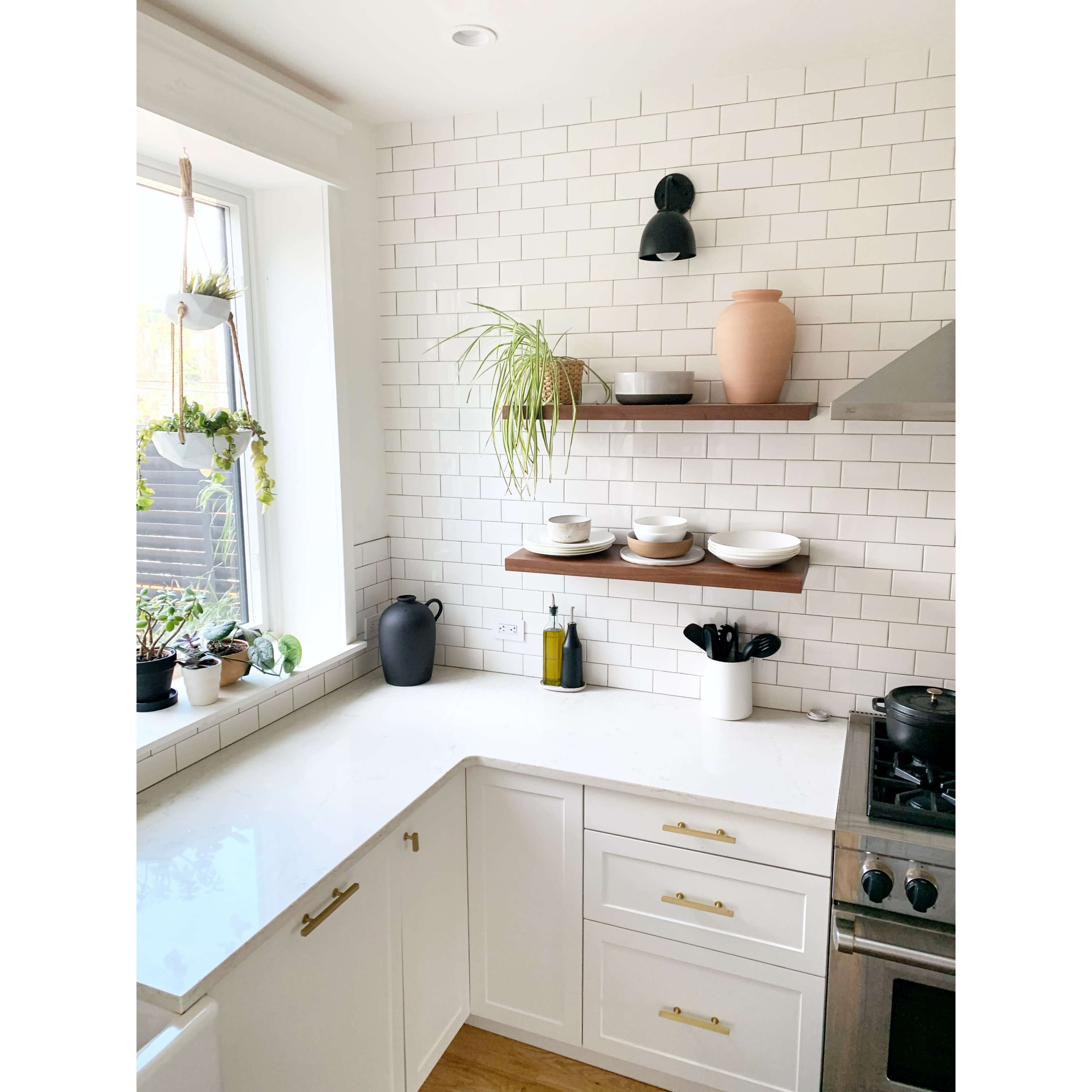 A modern kitchen features white subway tiles, floating wooden shelves with dishes and plants, and a corner sink with a view.