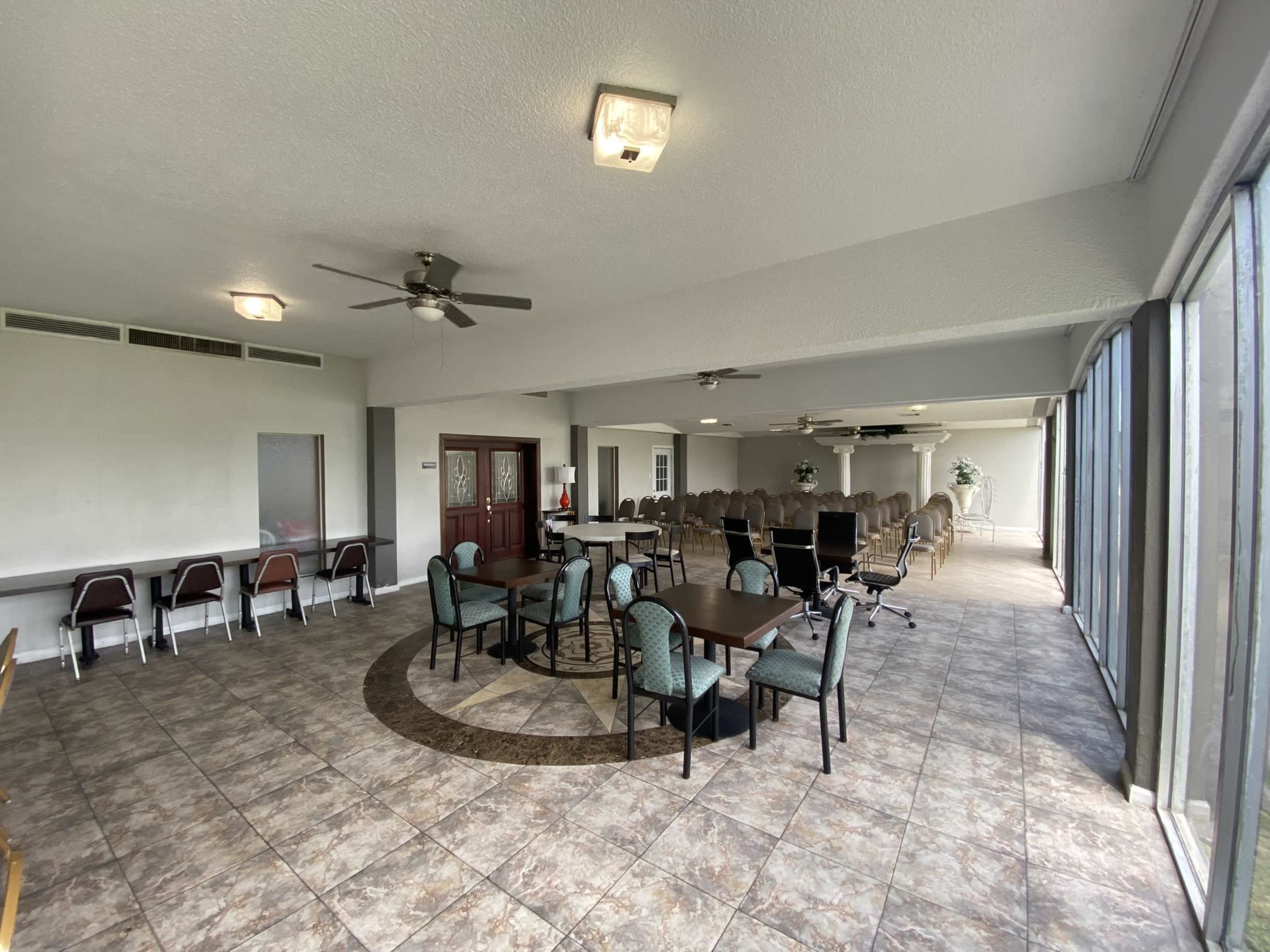 A spacious indoor area with tiled flooring, featuring a seating arrangement of tables and chairs on one side and a row of chairs set up in a theater-style layout on the other.