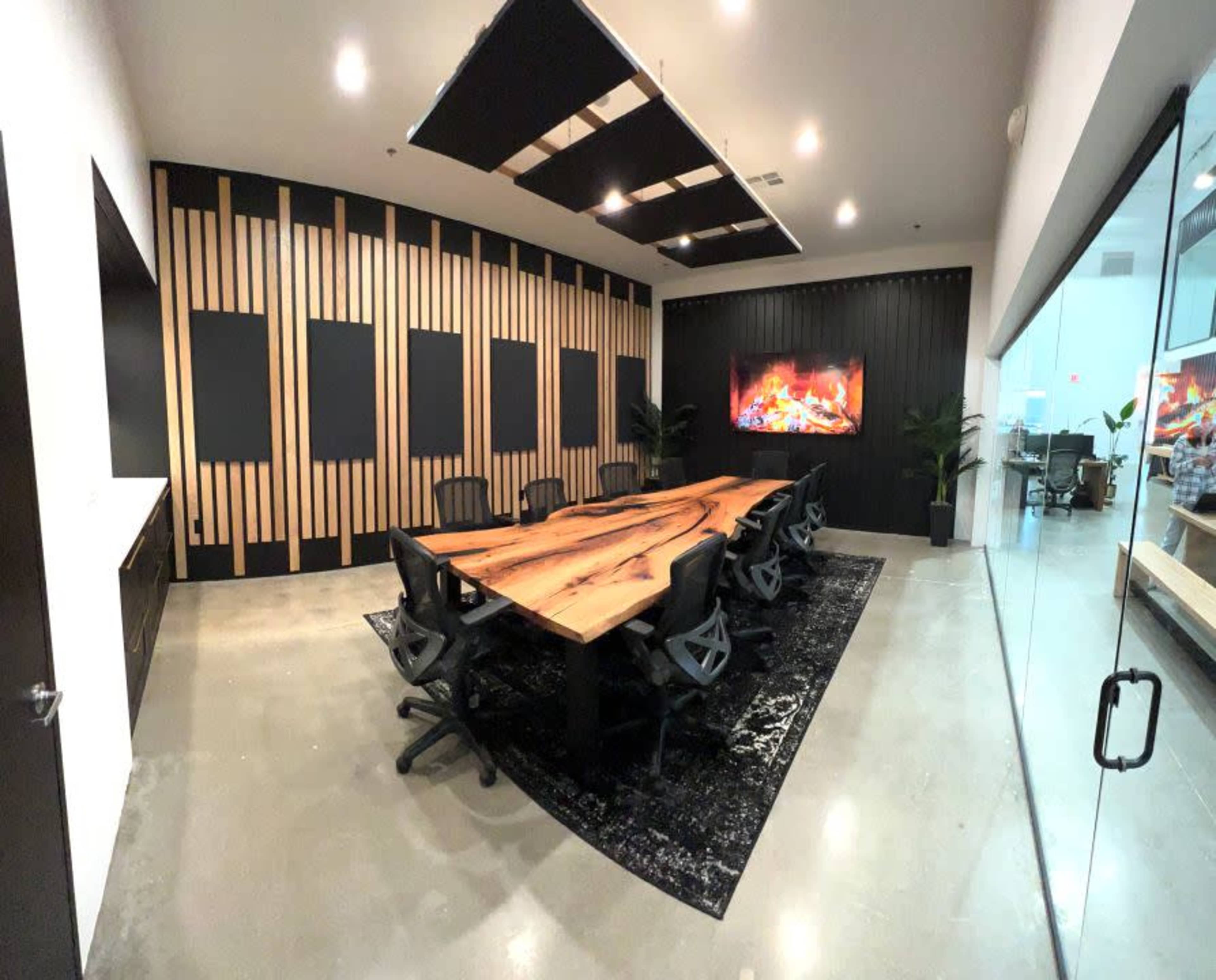 A modern conference room features a large wooden table surrounded by black chairs, with a textured wall and a digital fireplace display.